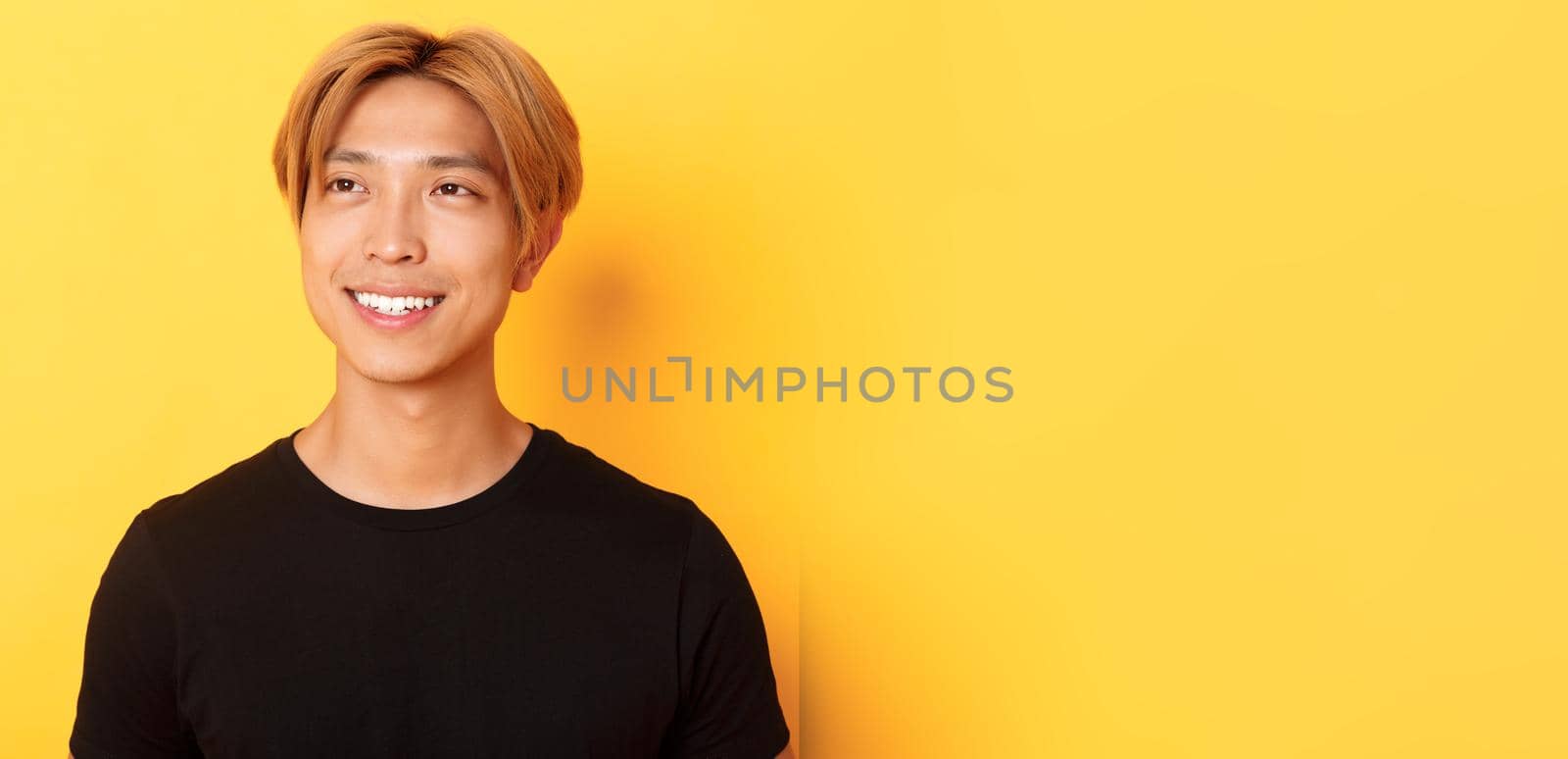 Close-up of handsome stylish korean guy with fair hair, looking dreamy and hopeful upper left corner, smiling pleased, standing yellow background.