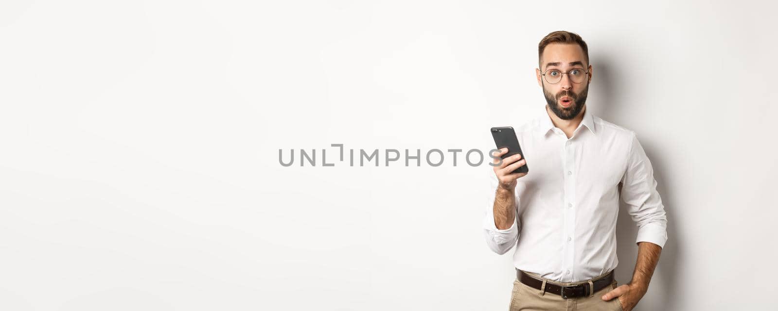 Surprised handsome man reading interesting info online, holding smartphone, standing over white background by Benzoix