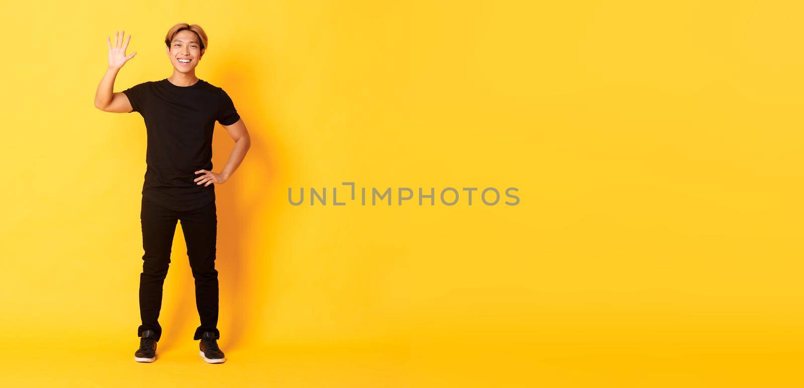 Full-length of friendly handsome asian man waving hand in hello, smiling and saying hi over yellow background by Benzoix