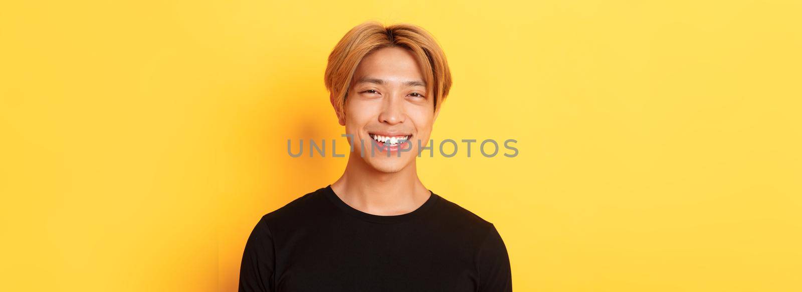 Close-up of handsome stylish asian guy with fair hair, smiling happy over yellow background.