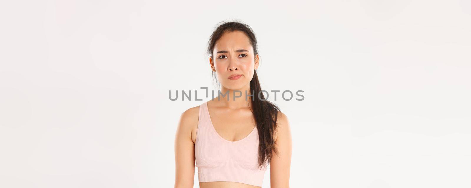 Sport, wellbeing and active lifestyle concept. Skeptical and displeased asian sportswoman, female athlete smirk disappointed and unamused, looking left with frowning face, white background.