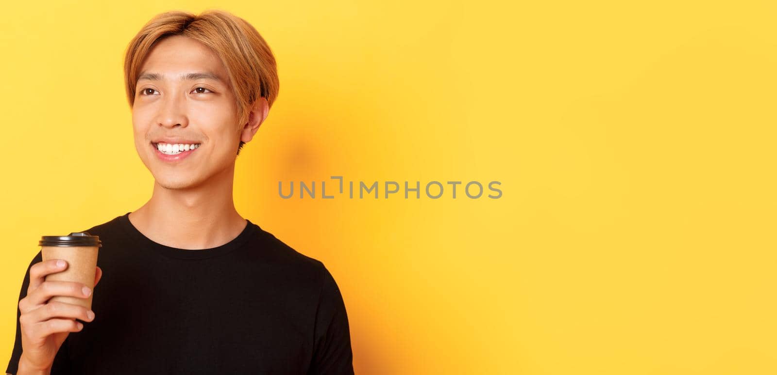 Close-up of relaxed and happy stylish asian blond guy, smiling and looking left dreamy while drinking coffee from takeaway cup, standing over yellow background by Benzoix