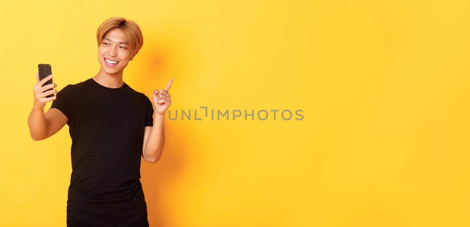 Portrait of handsome smiling asian man having video call on smartphone and pointing finger at something, standing yellow background.
