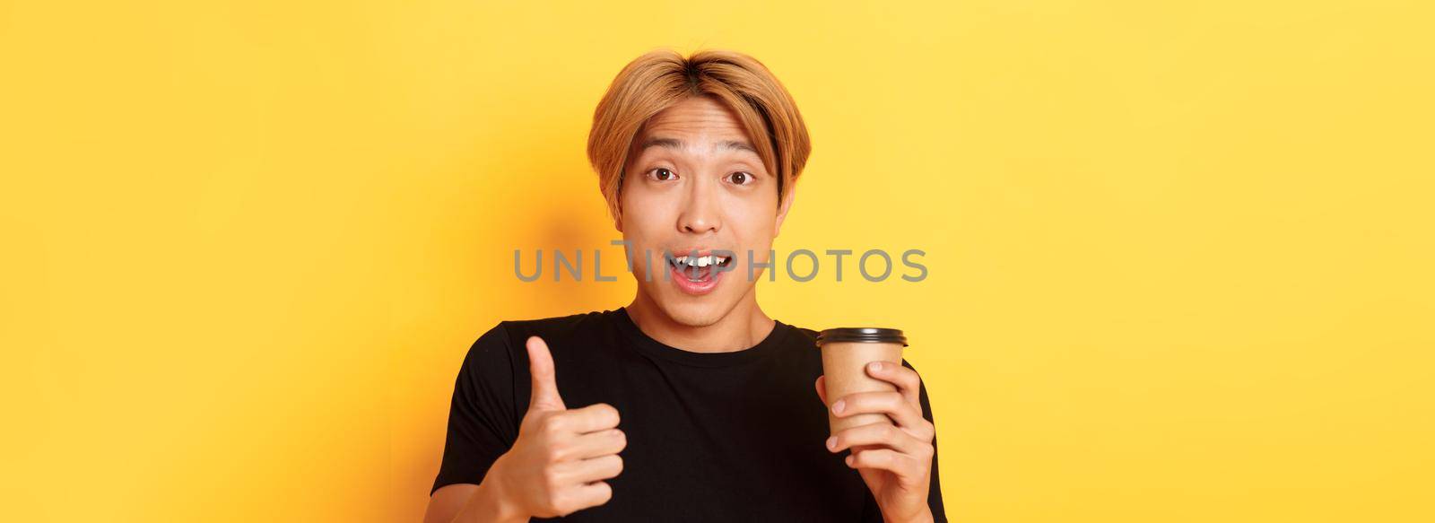 Close-up of surprised asian handsome guy recommend cafe, holding cup of coffee and showing thumbs-up in approval, smiling pleased over yellow background.