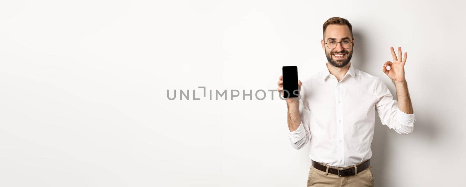 Satisfied young manager showing smartphone screen and okay sign, recommending application, standing over white background.