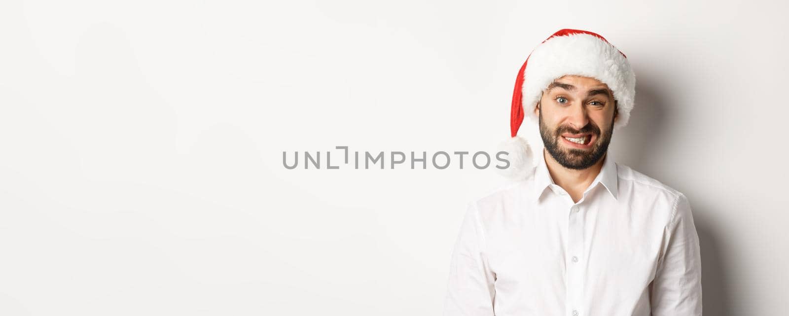 Close-up of awkward guy in santa hat apologizing, feeling uncomfortable, standing over white background. Christmas concept.