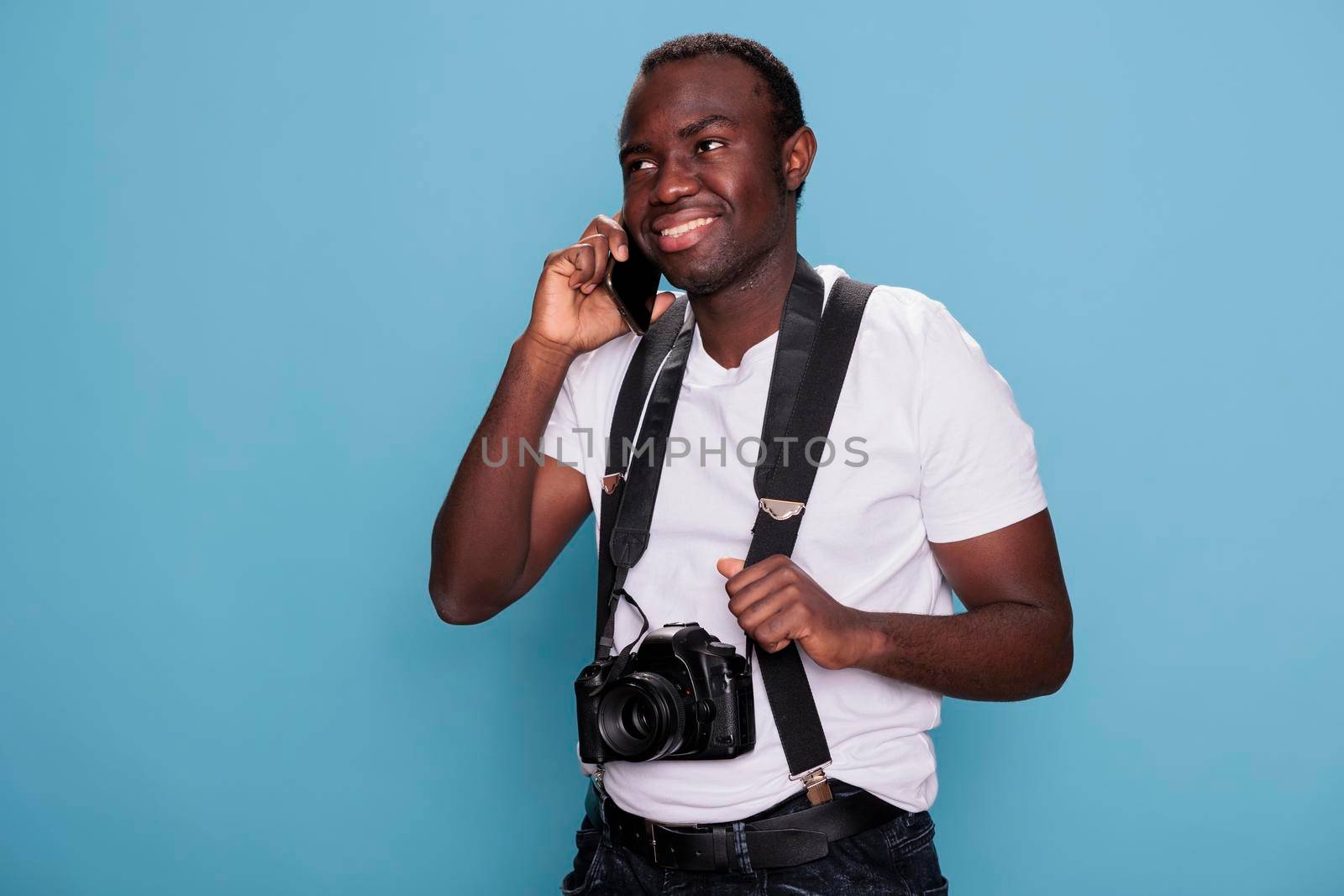Confident young photography enthusiast with camera speaking on modern touchscreen phone device.. Smiling professional photographer talking on smartphone while standing on blue background.