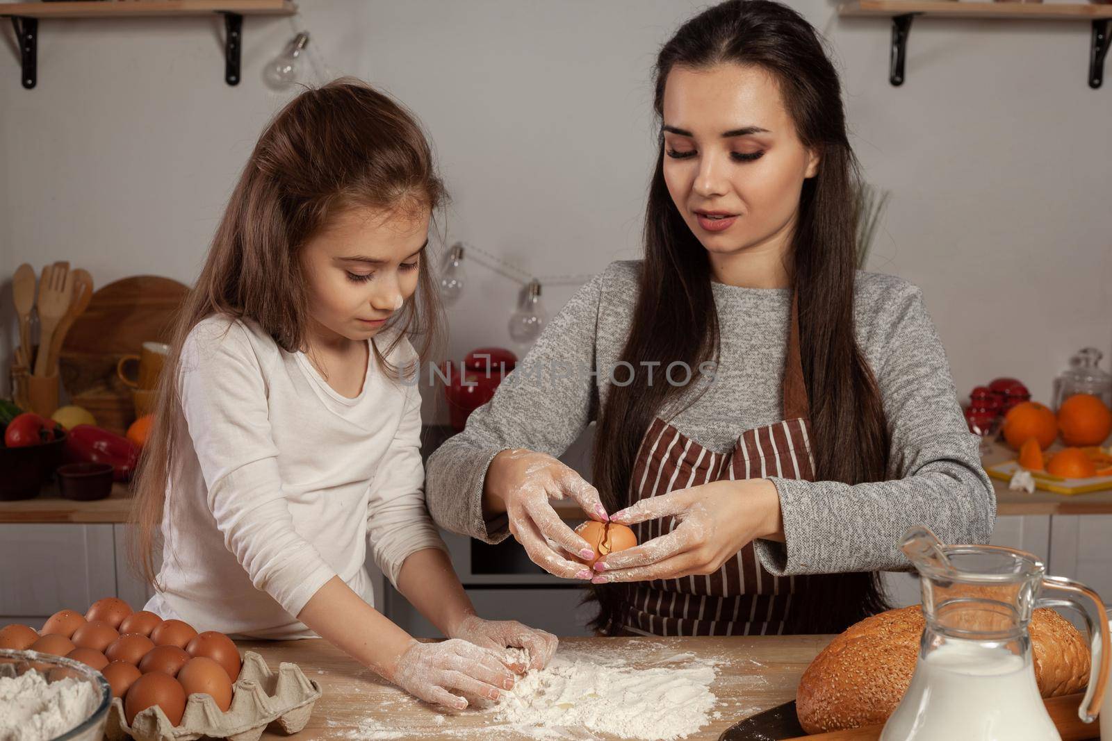 Happy loving family are preparing pastries together. Cute mommy and her little girl are adding an egg to the dough and having fun at the kitchen, against a white wall with shelves and bulbs on it. Homemade food and little helper.