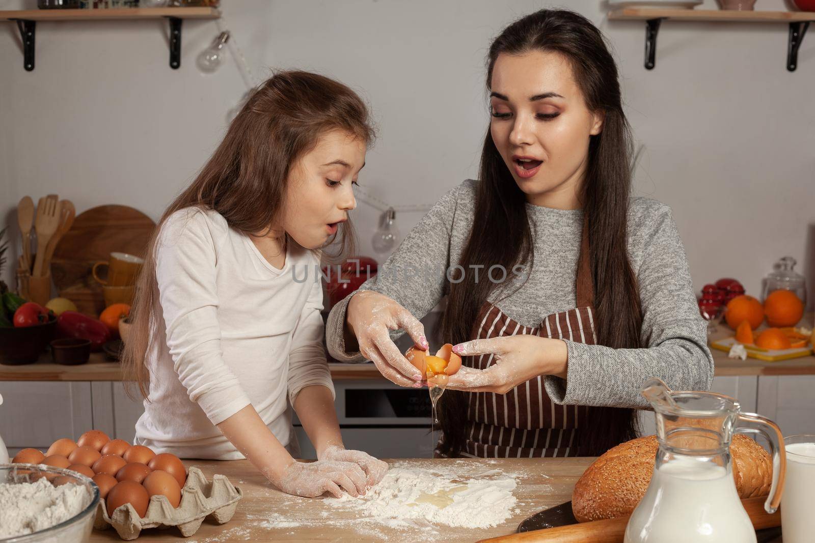 Happy loving family are preparing pastries together. Cute mother and her daughter are adding an egg to the dough and having fun at the kitchen, against a white wall with shelves and bulbs on it. Homemade food and little helper.