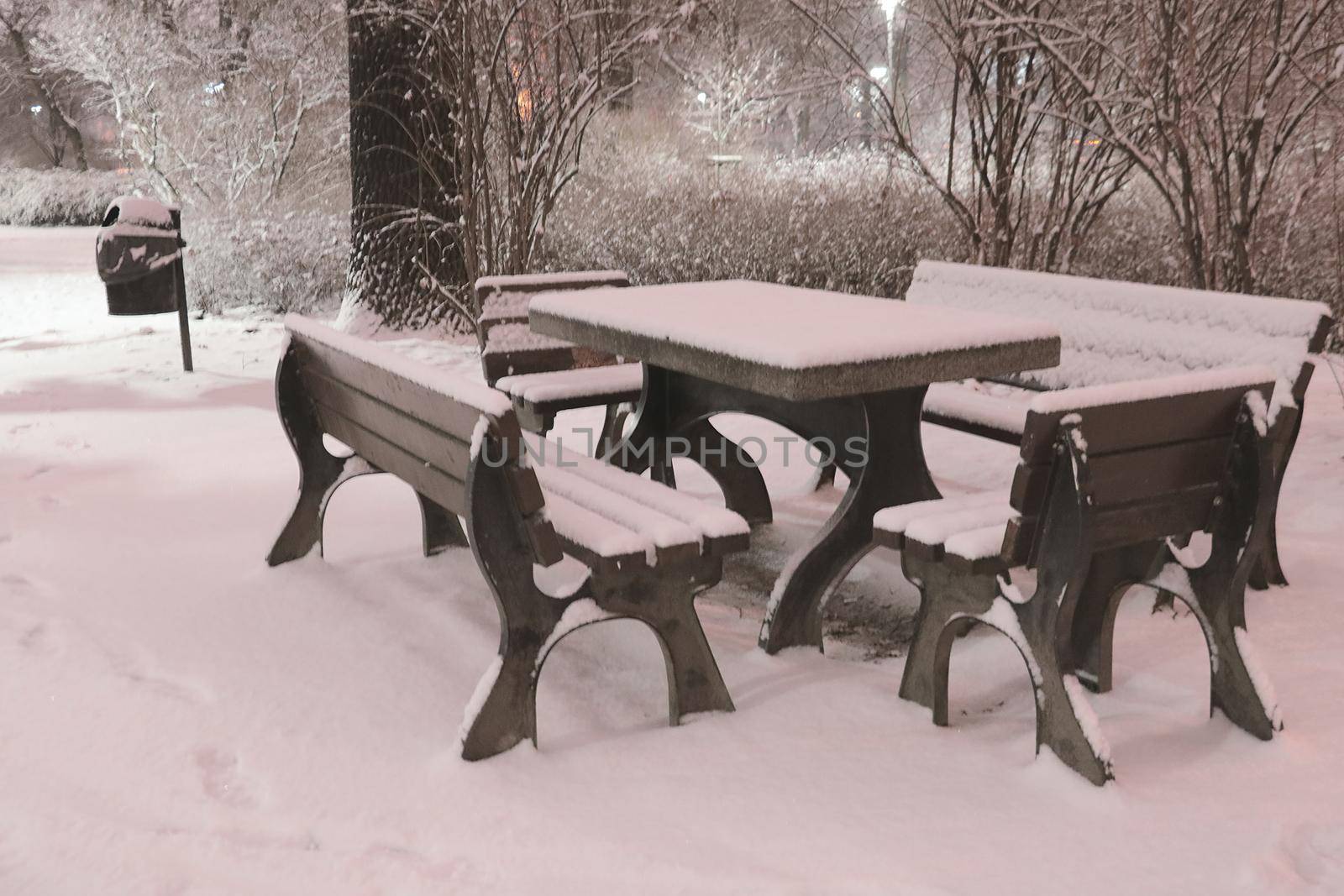 Black and white photo. Snow-covered benches and a table in the park in the evening