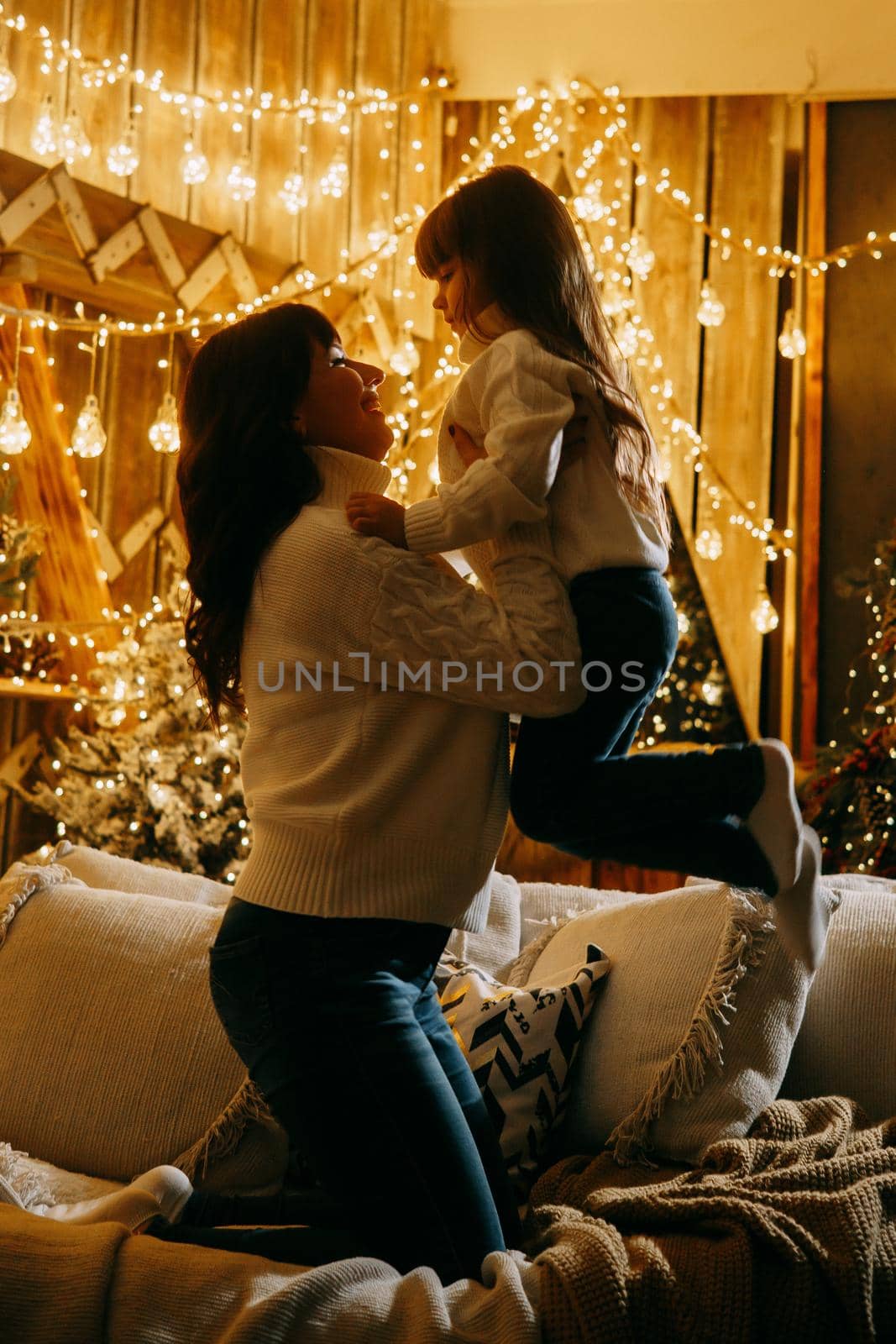 A little girl with her mother in a cozy home environment on the sofa next to the Christmas tree. The theme of New Year holidays and festive interior with garlands and light bulbs. by Annu1tochka