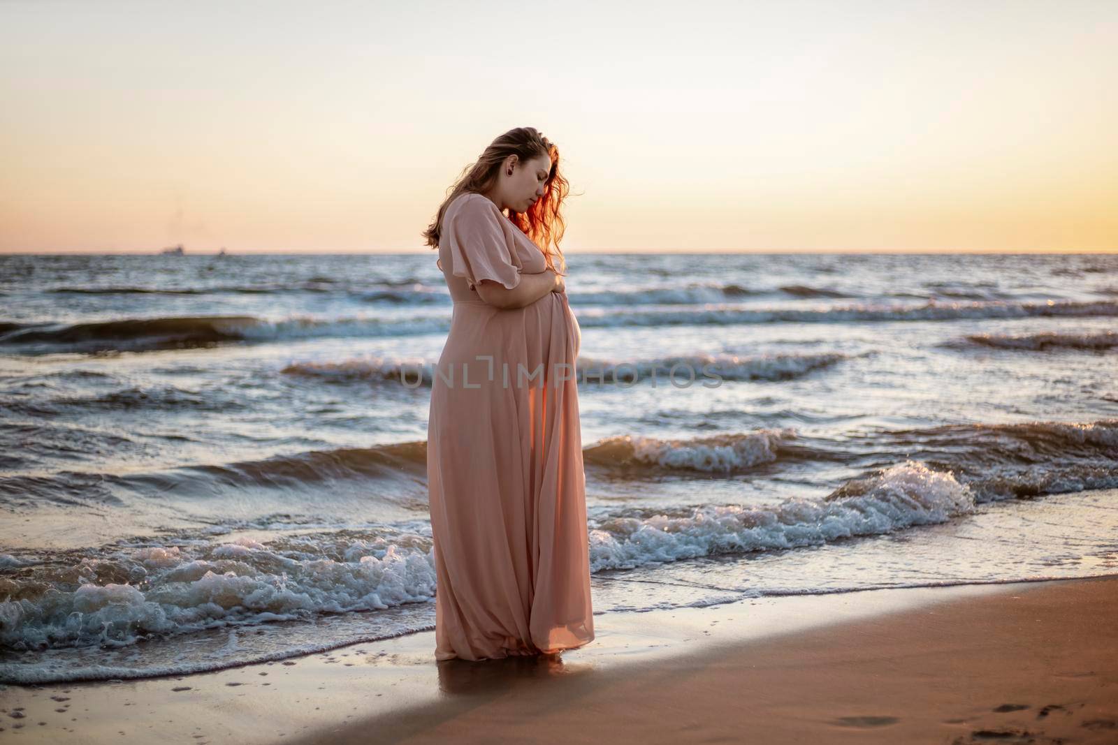 Young pregnant woman with a beautiful sea view on the background. Happy and calm pregnant woman with long hair and pink dress standig on the beach. Romantic view, ocean, sunset, maternity. by creativebird