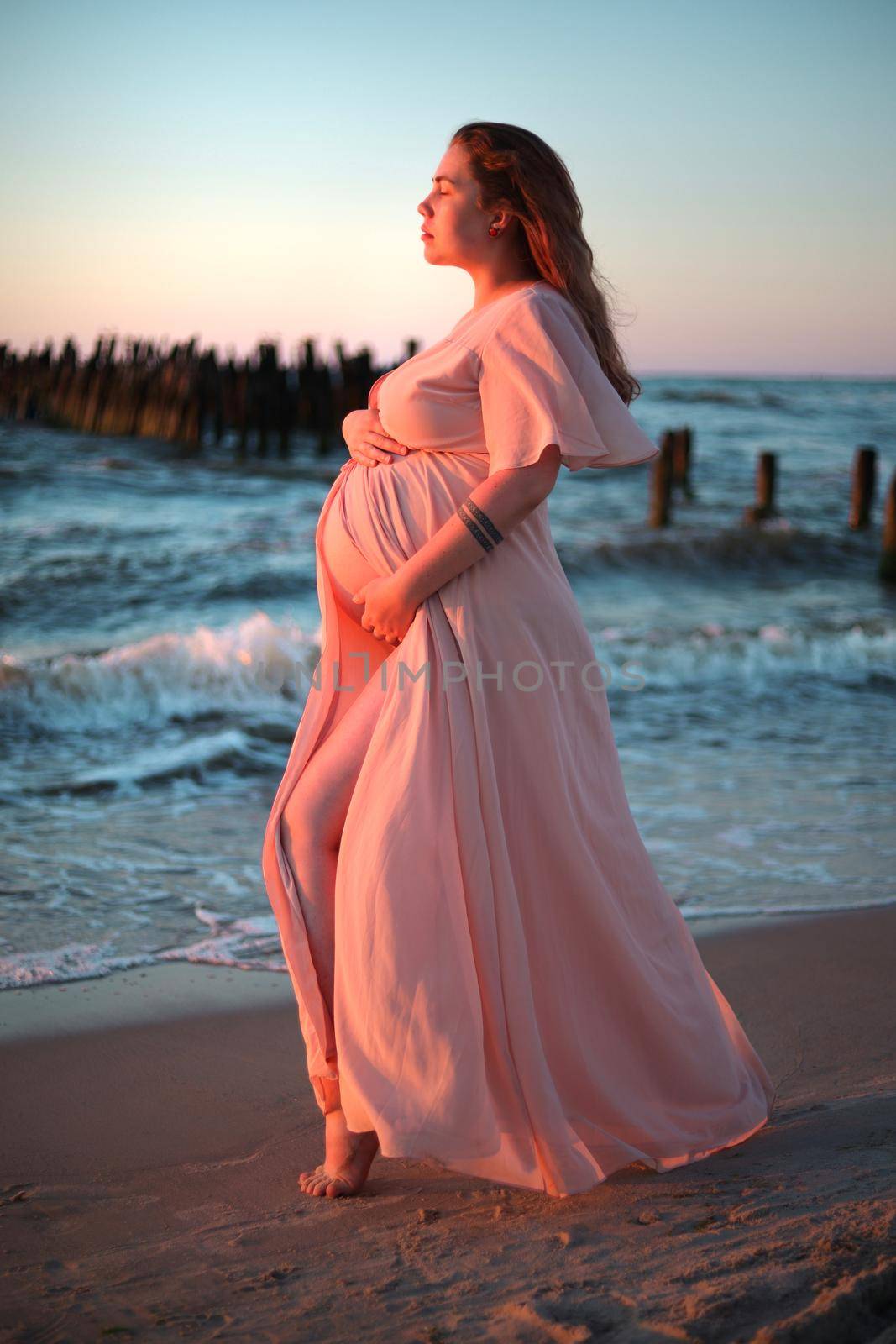 Young pregnant woman with a beautiful sea view on the background. Happy and calm pregnant woman with long hair and pink dress standig on the beach. Maternity. by creativebird