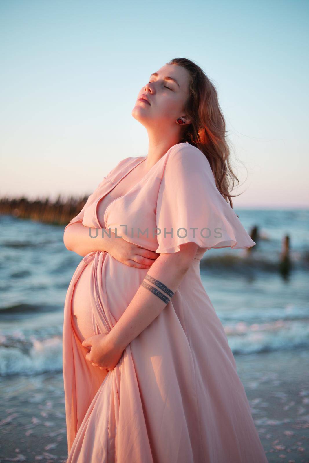 Young pregnant woman with a beautiful sea view on the background. Happy and calm pregnant woman with long hair and pink dress standig on the beach. Romantic view, ocean, sunset, maternity