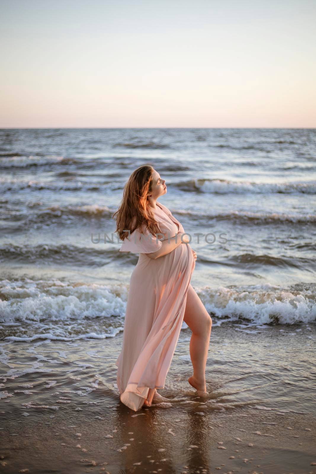 Young pregnant woman with a beautiful sea view on the background. Happy and calm pregnant woman with long hair and pink dress standig on the beach. Romantic view, ocean, sunset, maternity