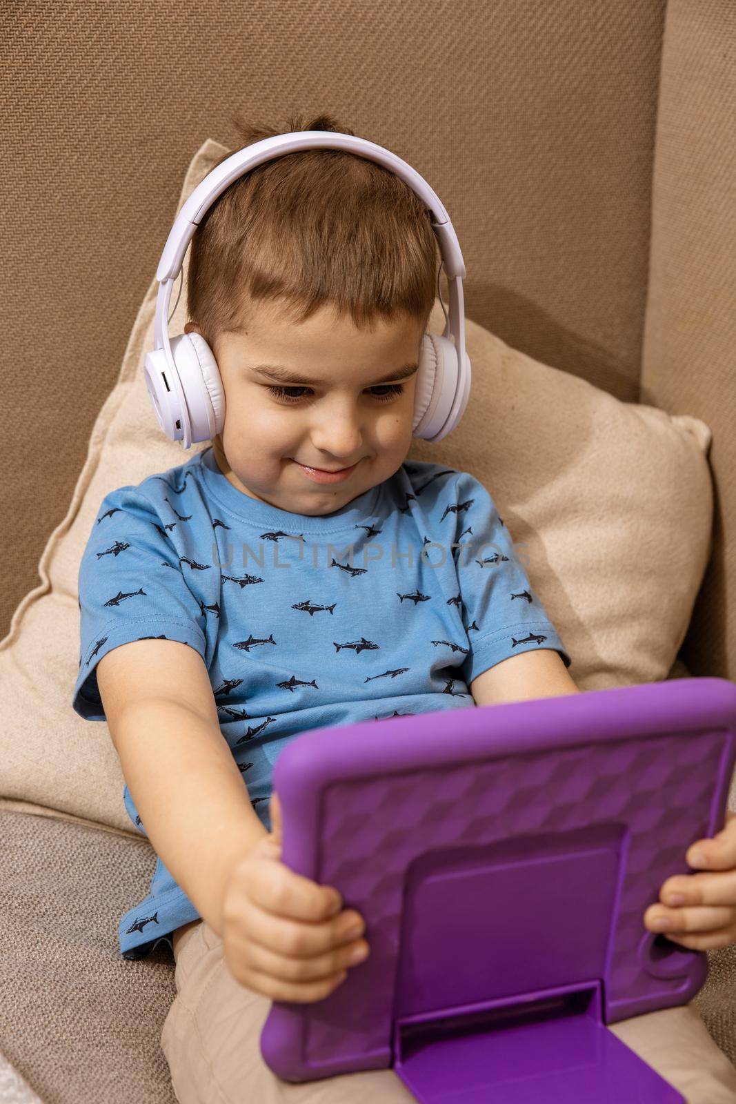 Little caucasian boy with blue shirt and white headphones watching cartoon on violet digital tablet at home. Modern child and education technology. by creativebird