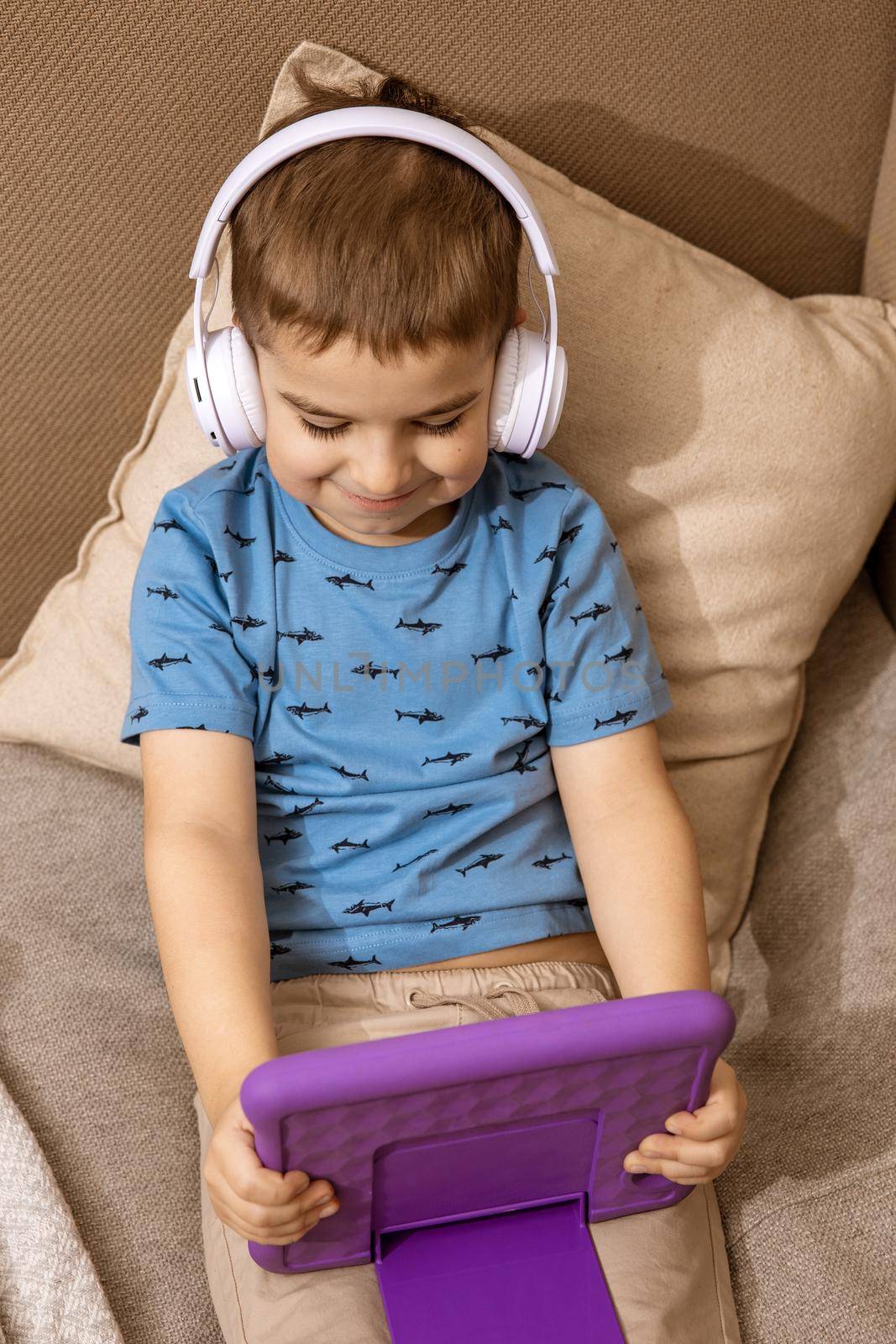 Little caucasian boy with blue shirt and white headphones watching cartoon on violet digital tablet at home. Modern child and education technology