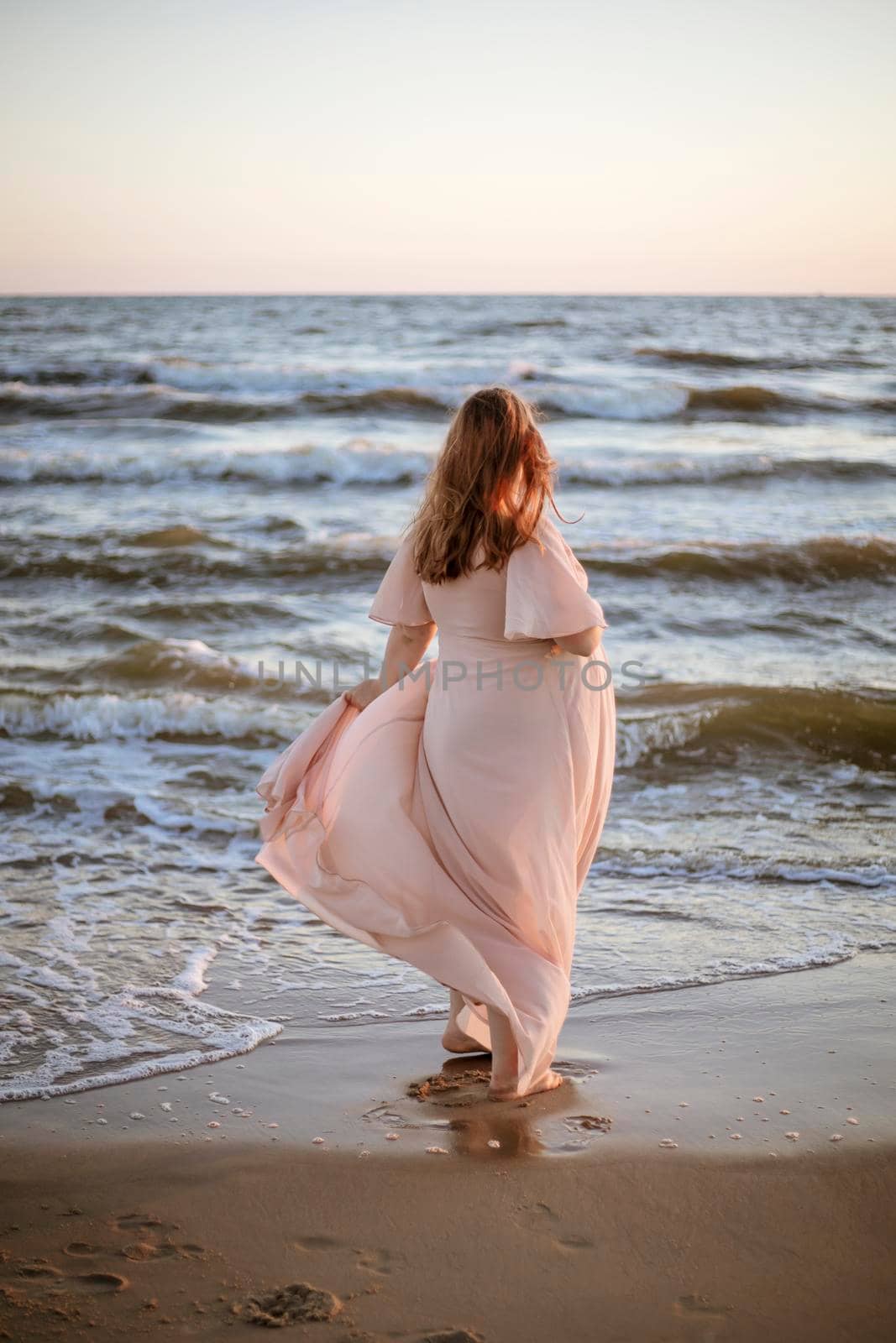 Young woman with a beautiful sea view on the background. Woman with long hair and pink dress standig on the beach. Romantic view, ocean, sunset, relax. Sekective focus