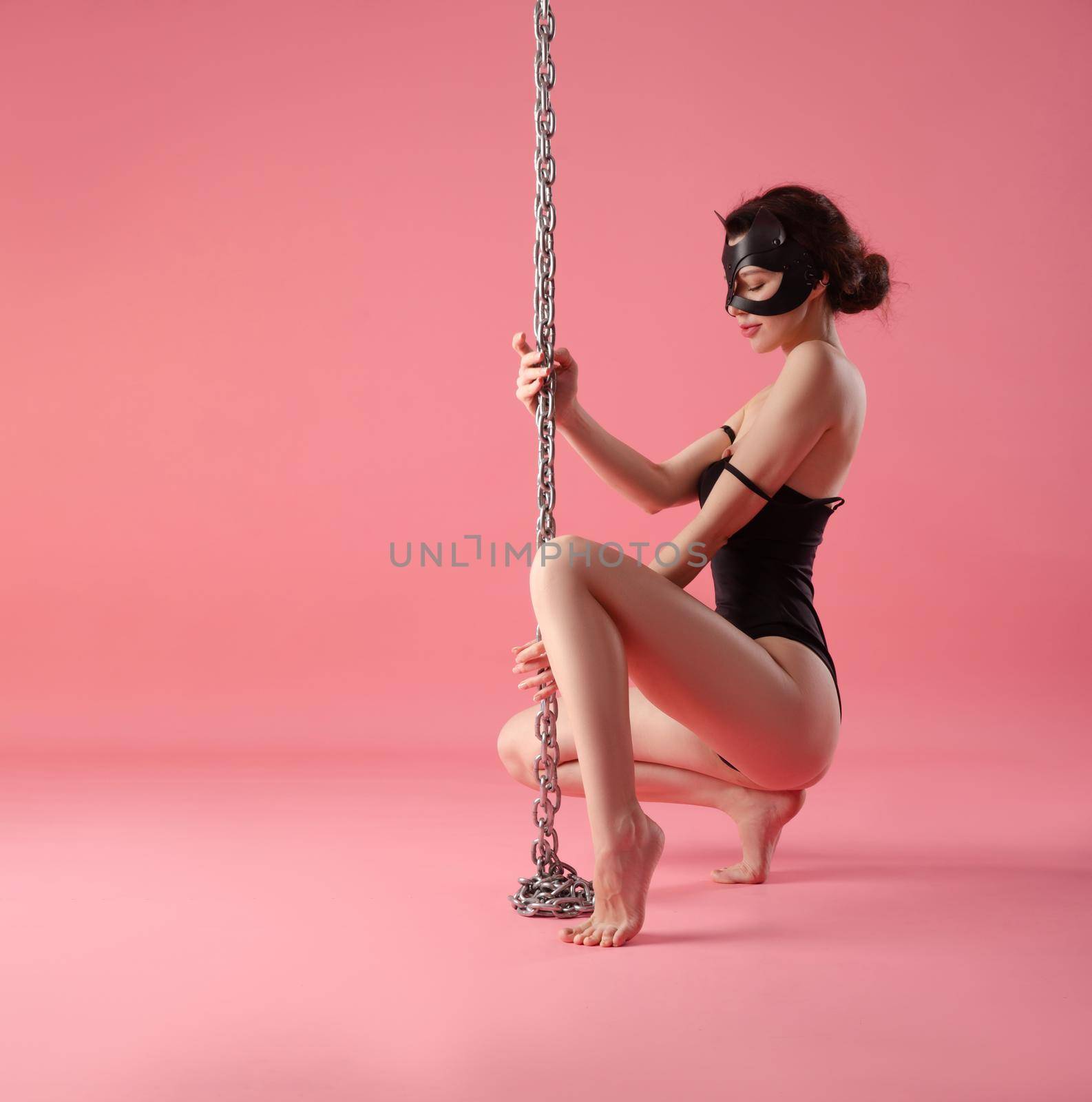 girl in a bdsm cat mask with a metal chain and leather bracelets on her feet on the floor on a pink background