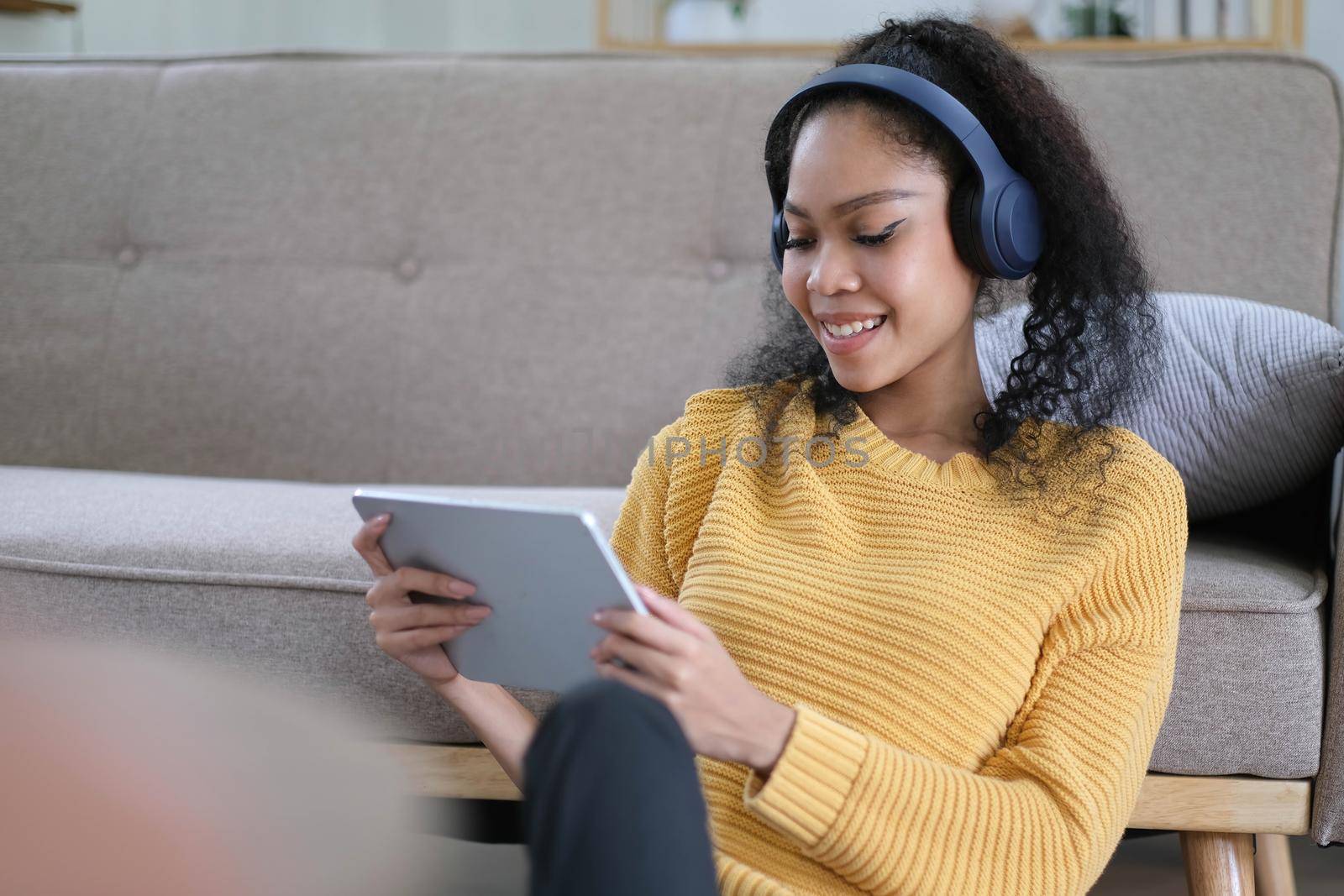 Photo of beautiful dark skin wavy lady homey mood holding digital tablet earflaps listening new audio sitting floor near couch casual clothes yellow at living room indoors by wichayada