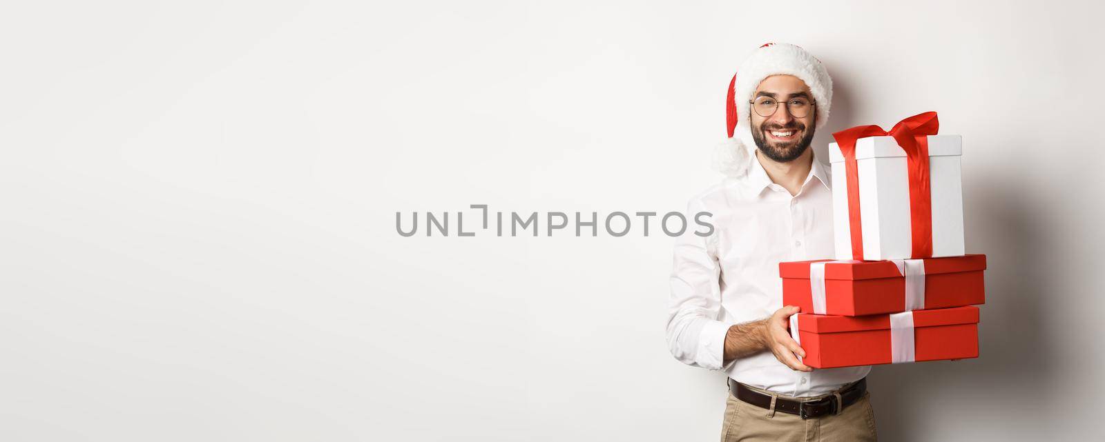 Merry christmas, holidays concept. Happy young man smiling, holding gifts in boxes and wearing santa hat, white background.