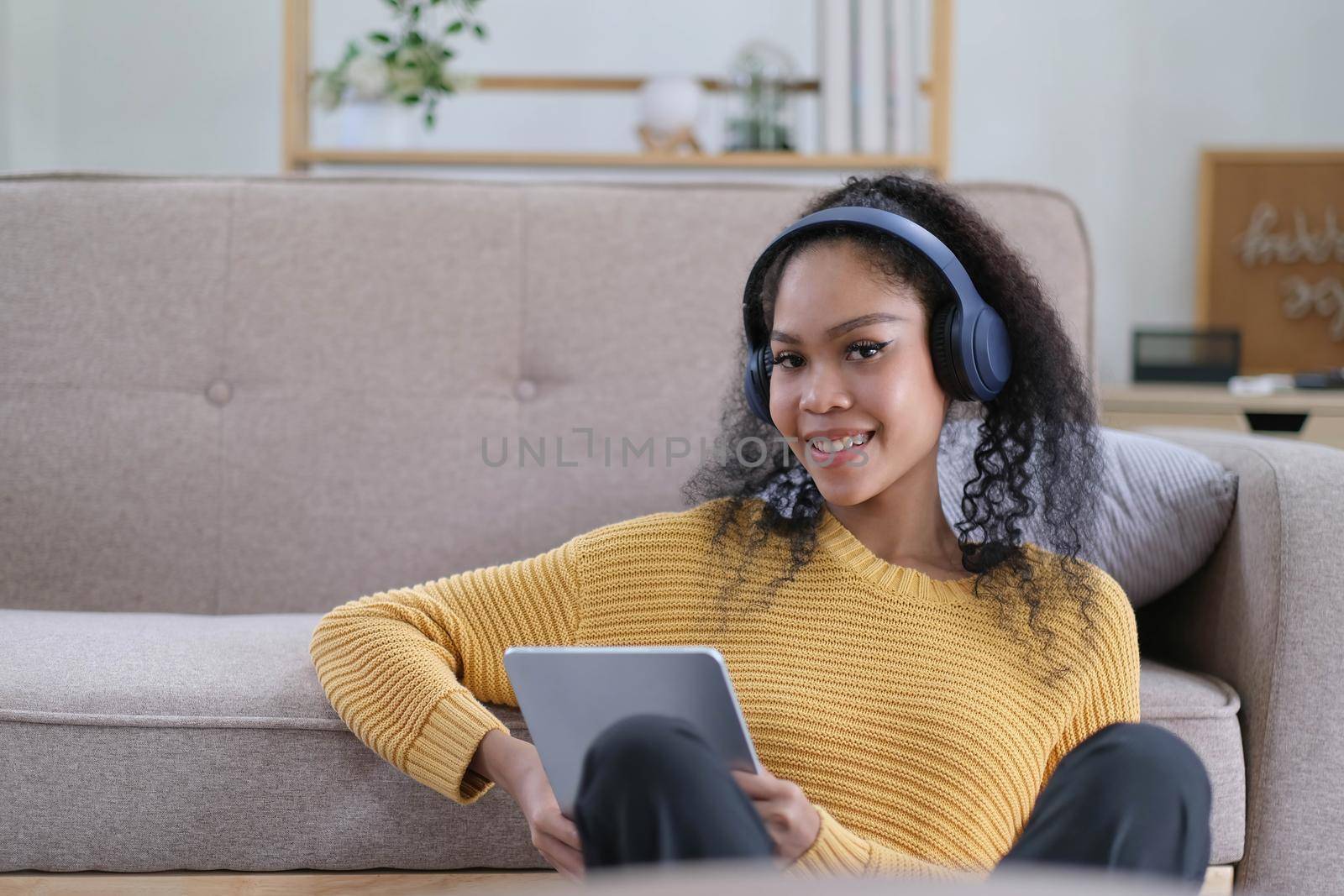 Photo of beautiful dark skin wavy lady homey mood holding digital tablet earflaps listening new audio sitting floor near couch casual clothes yellow at living room indoors by wichayada