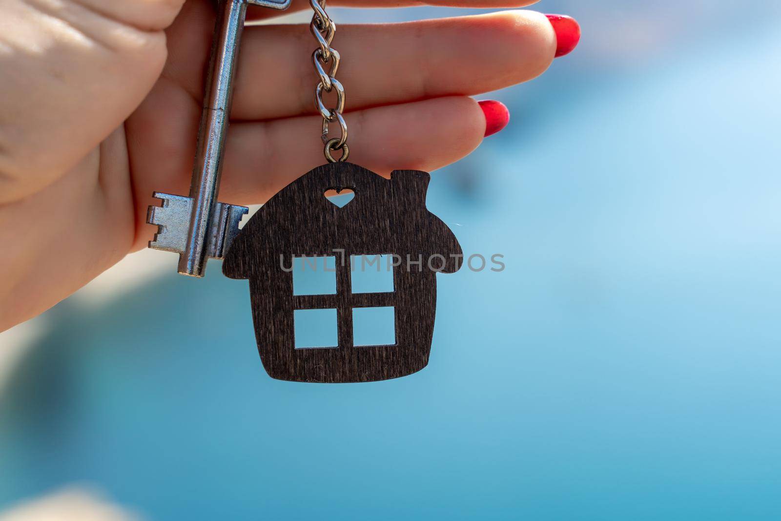 Keys with a keychain in the shape of a house in a female hand against the backdrop of the sea. The concept of buying a house, apartment, real estate, young family, mortgage loan, rental housing. by Matiunina