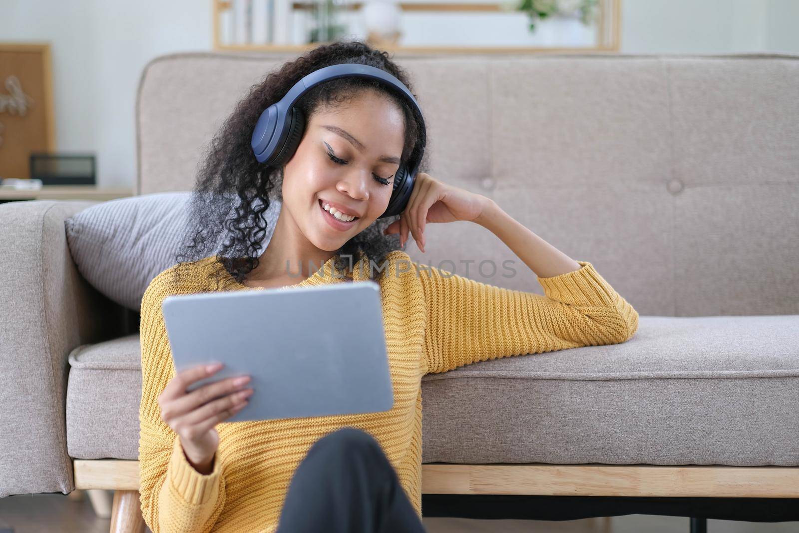 Photo of beautiful dark skin wavy lady homey mood holding digital tablet earflaps listening new audio sitting floor near couch casual clothes yellow at