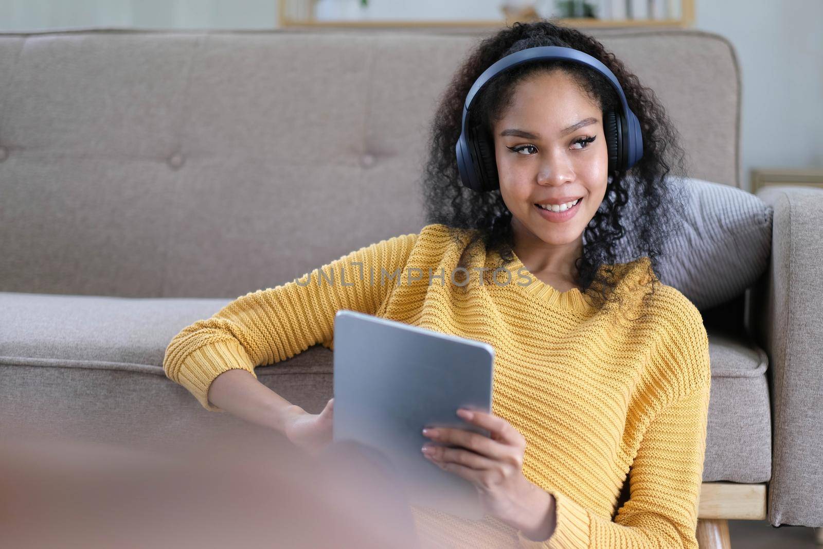 Photo of beautiful dark skin wavy lady homey mood holding digital tablet earflaps listening new audio sitting floor near couch casual clothes yellow at living room indoors by wichayada