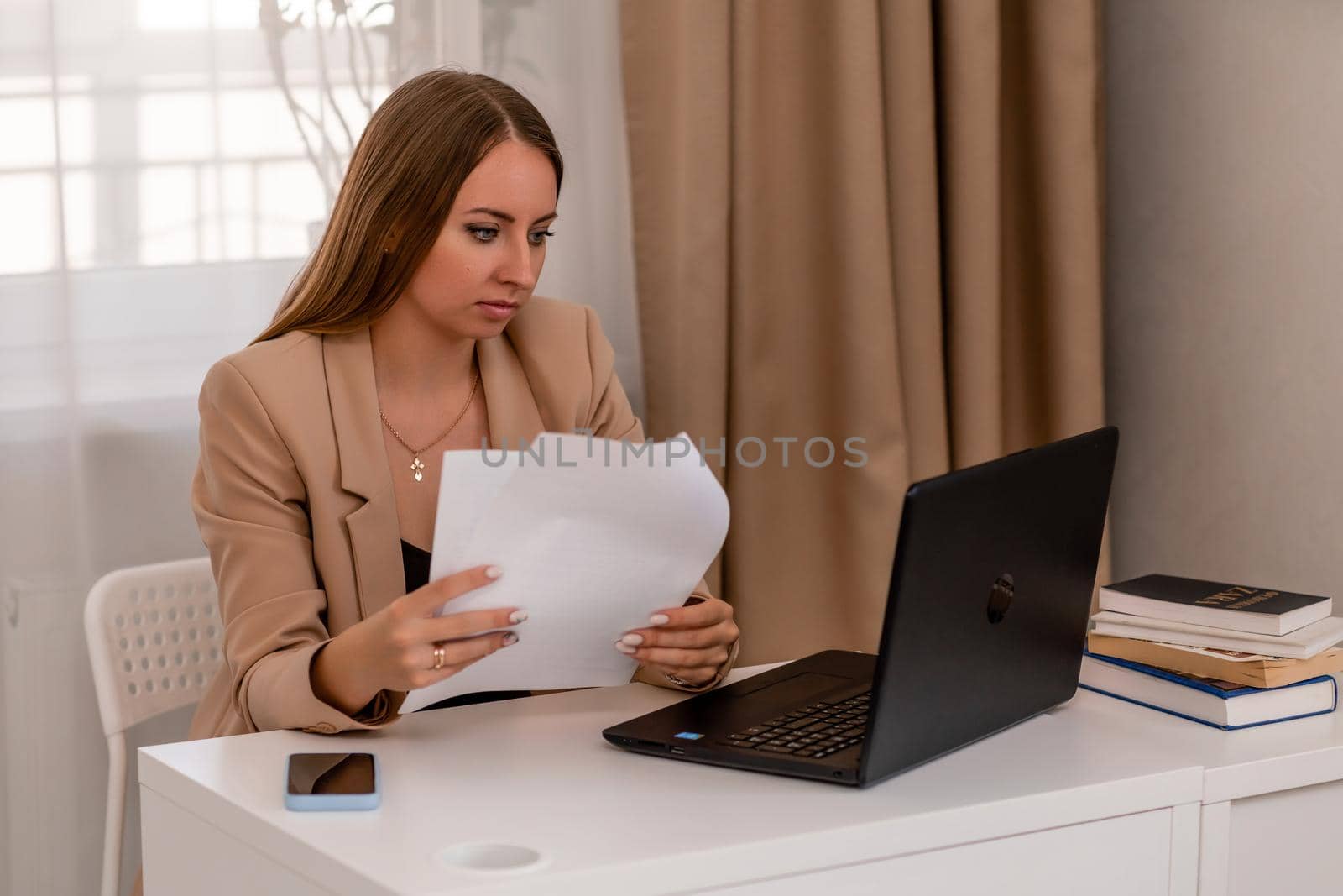 European professional woman sitting with laptop at home office desk, positive woman studying while working on PC. She is wearing a beige jacket and jeans. by Matiunina