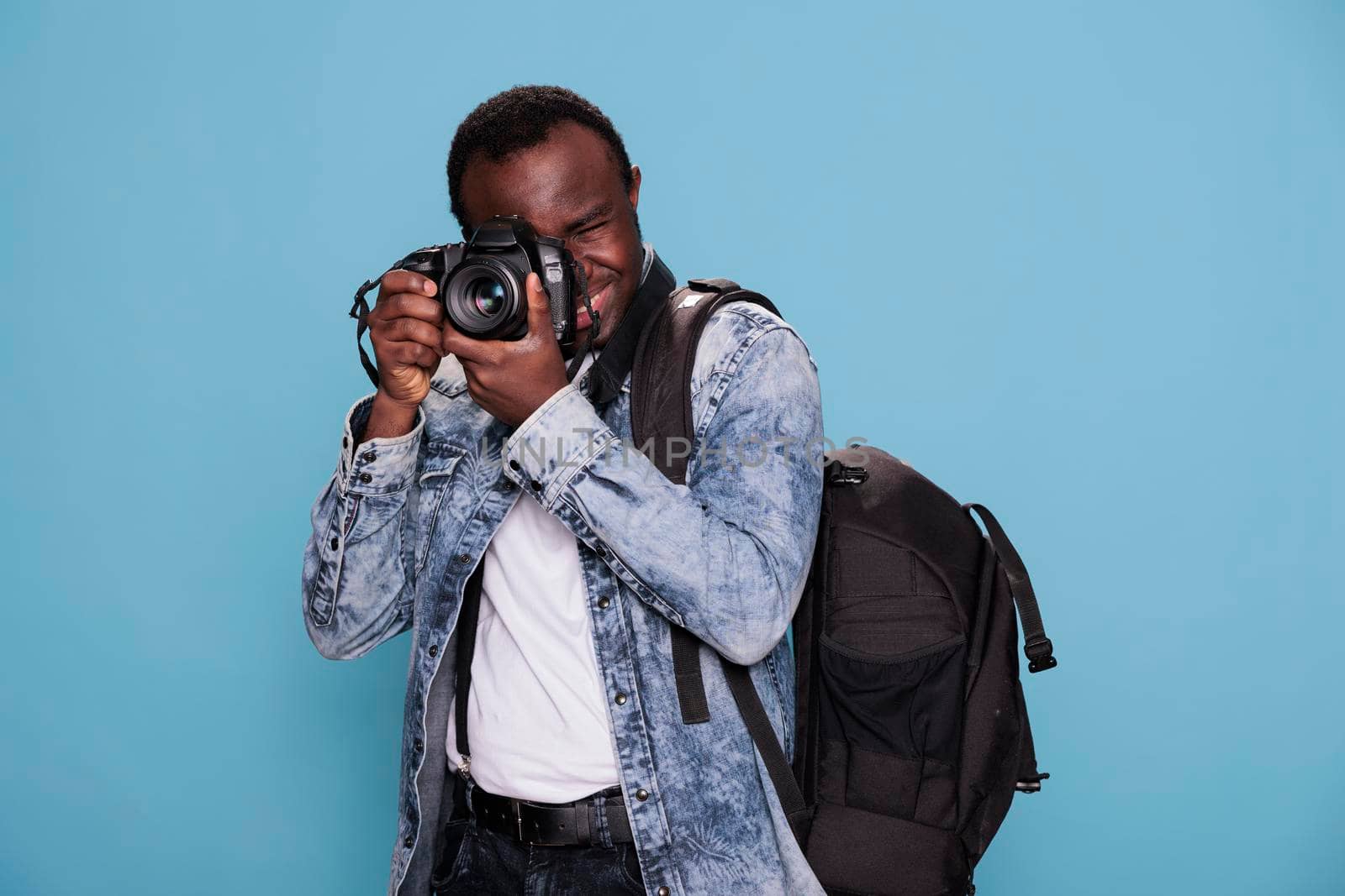 Young professional photographer with DSLR camera taking photo while standing on blue background. Confident photography enthusiast having photo device while taking picture.