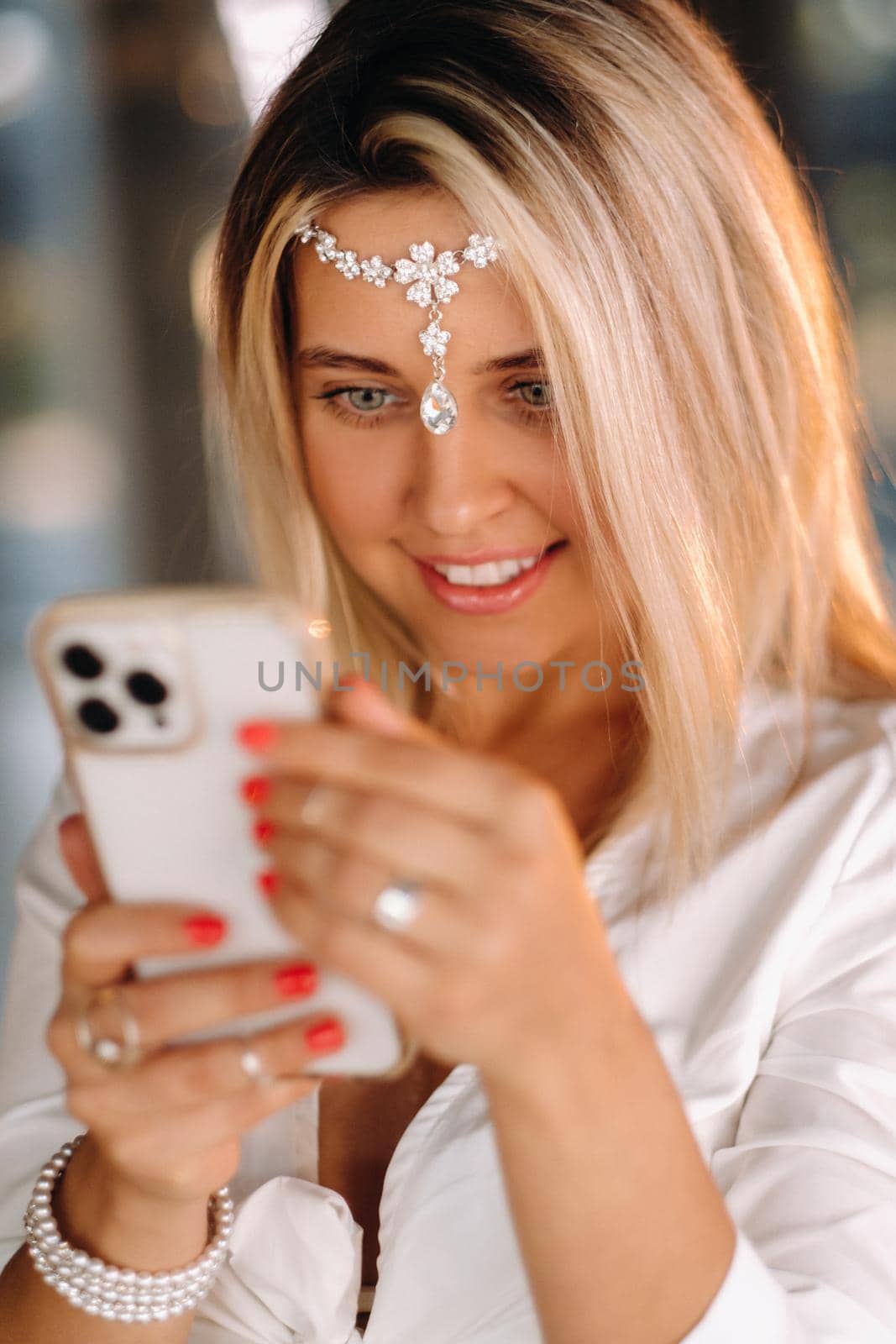 A positive young woman in a white dress and an ornament on her head was smiling, holding a phone in her hands and looking into it.