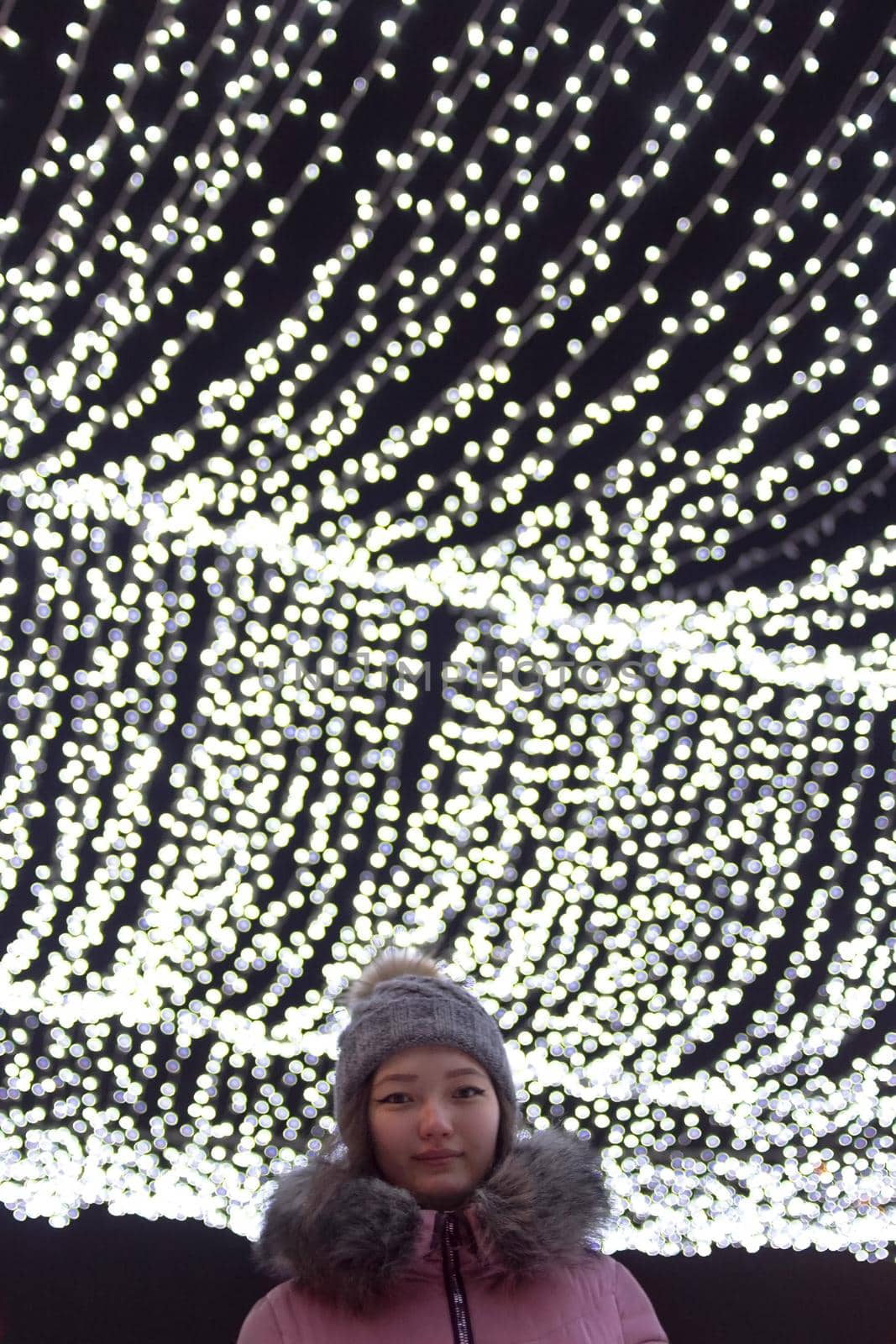 Low angle headshot of young woman with christmas lights on background at night, sky festive illumination shine and glow, urban holiday, vertical image with copy space