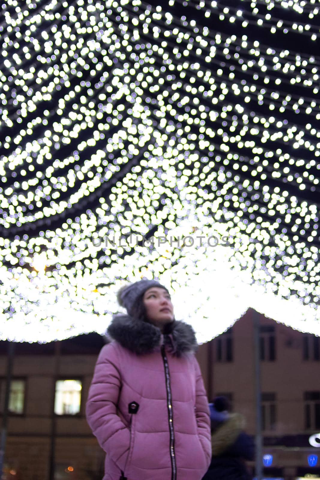 Young woman looks up, central downtown with christmas lights at night, sky festive illumination shine and glow, urban holiday, vertical defocus image with copy space, low angle