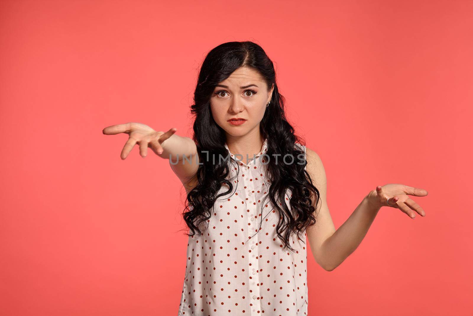 Studio shot of an attractive teen lady wearing casual white polka dot blouse. Little brunette female looking upset posing over a pink background. People and sincere emotions.