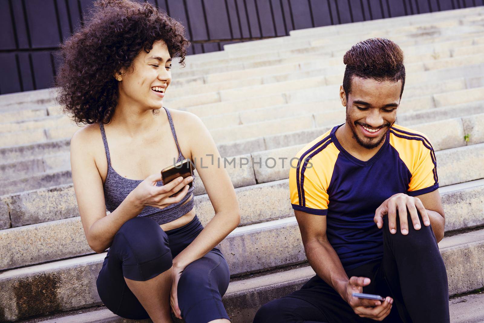 athletic latin woman smiles happy with a smiling black sports man and have fun with mobile phone on the stairs during a rest in the workout, technology in sport and healthy lifestyle concept