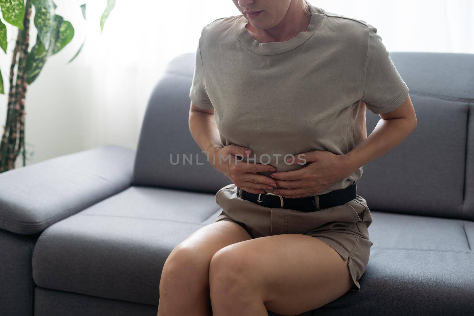 Sad, upset, unhappy woman holding hands on stomach suffering from abdominal pain with close eyes, having menstrual period, food poisoning, gastritis, diarrhea, feeling unwell sitting in livingroom. by Andelov13