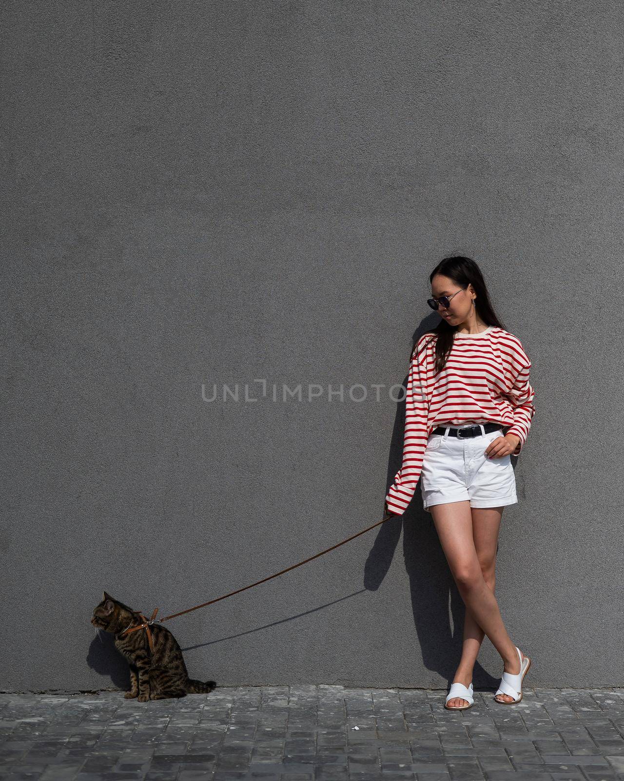 A young woman walks with a gray tabby cat on a leash against a gray wall. by mrwed54