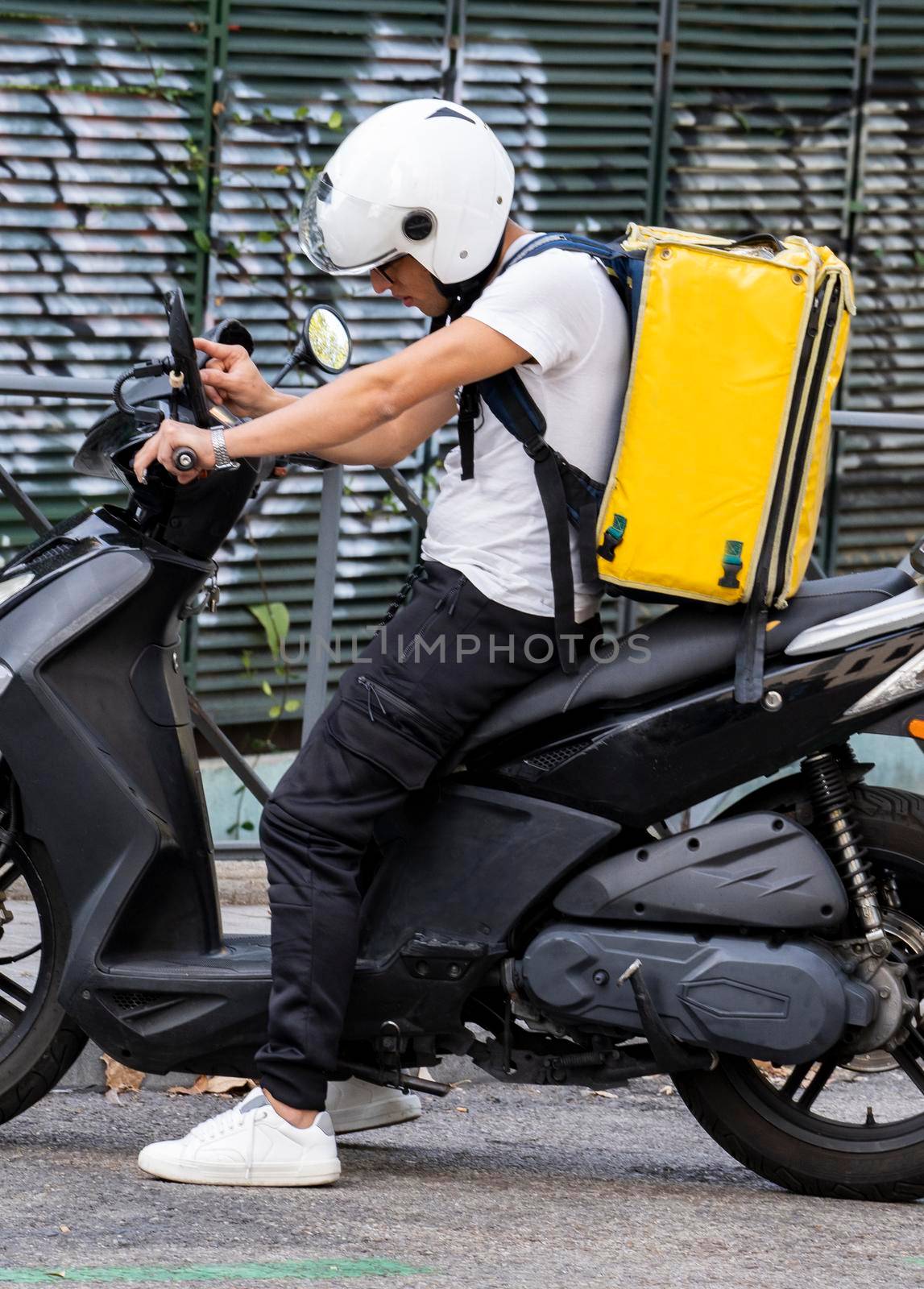 Young courier, delivery man searching his destination on the mobile , with thermo backpack on a moped .Online order. by papatonic