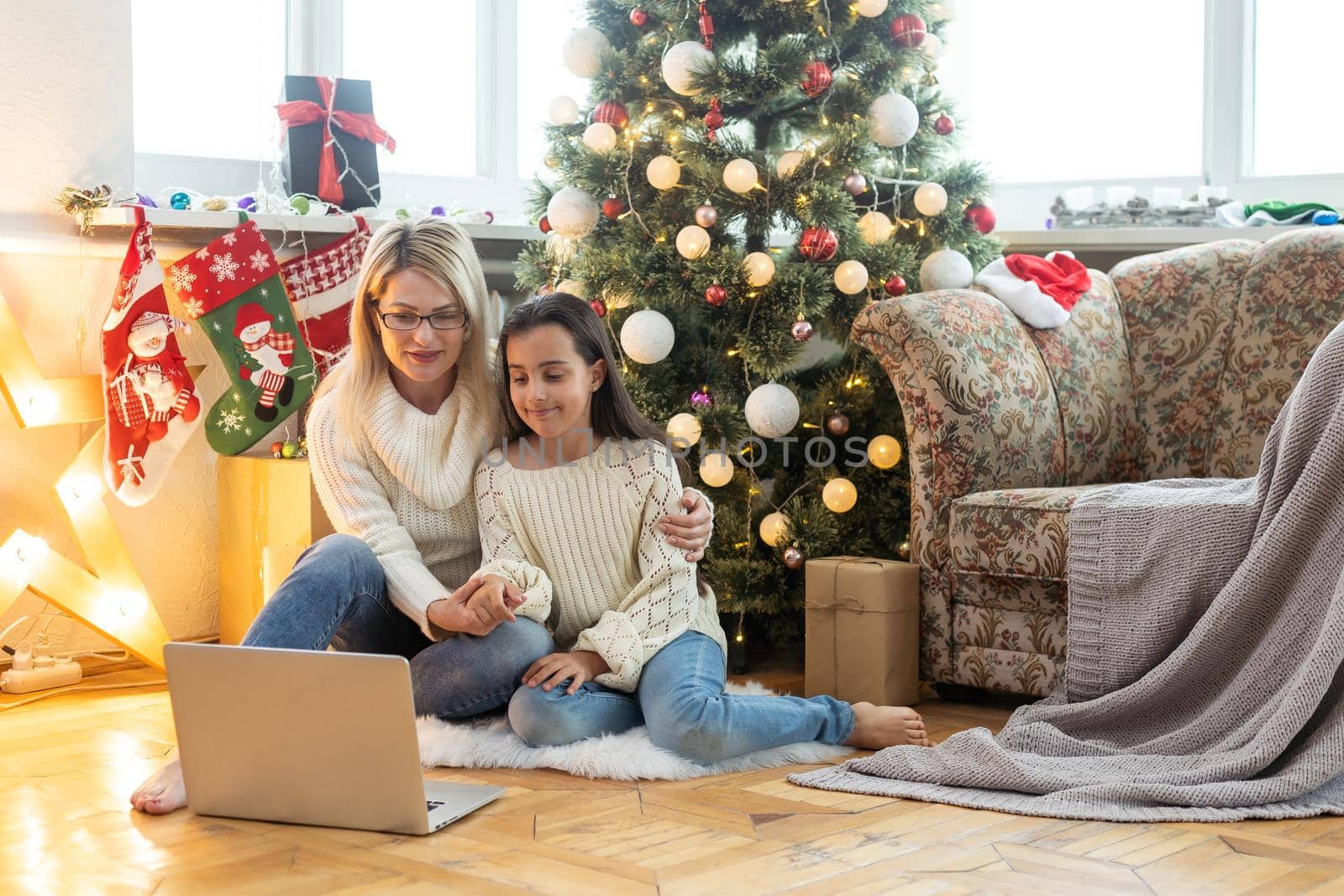 Indoor shot of beautiful happy young woman shopping online on laptop in cozy Christmas interior. Mother on the floor next the Christmas tree and sofa and daughter embrace her, shopping gifts