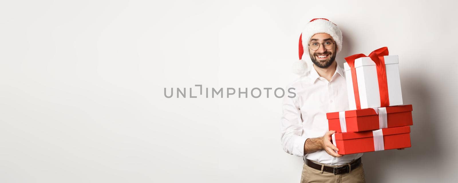 Winter holidays and celebration. Happy guy bring christmas presents, holding gifts and wearing Santa hat, standing over white background.