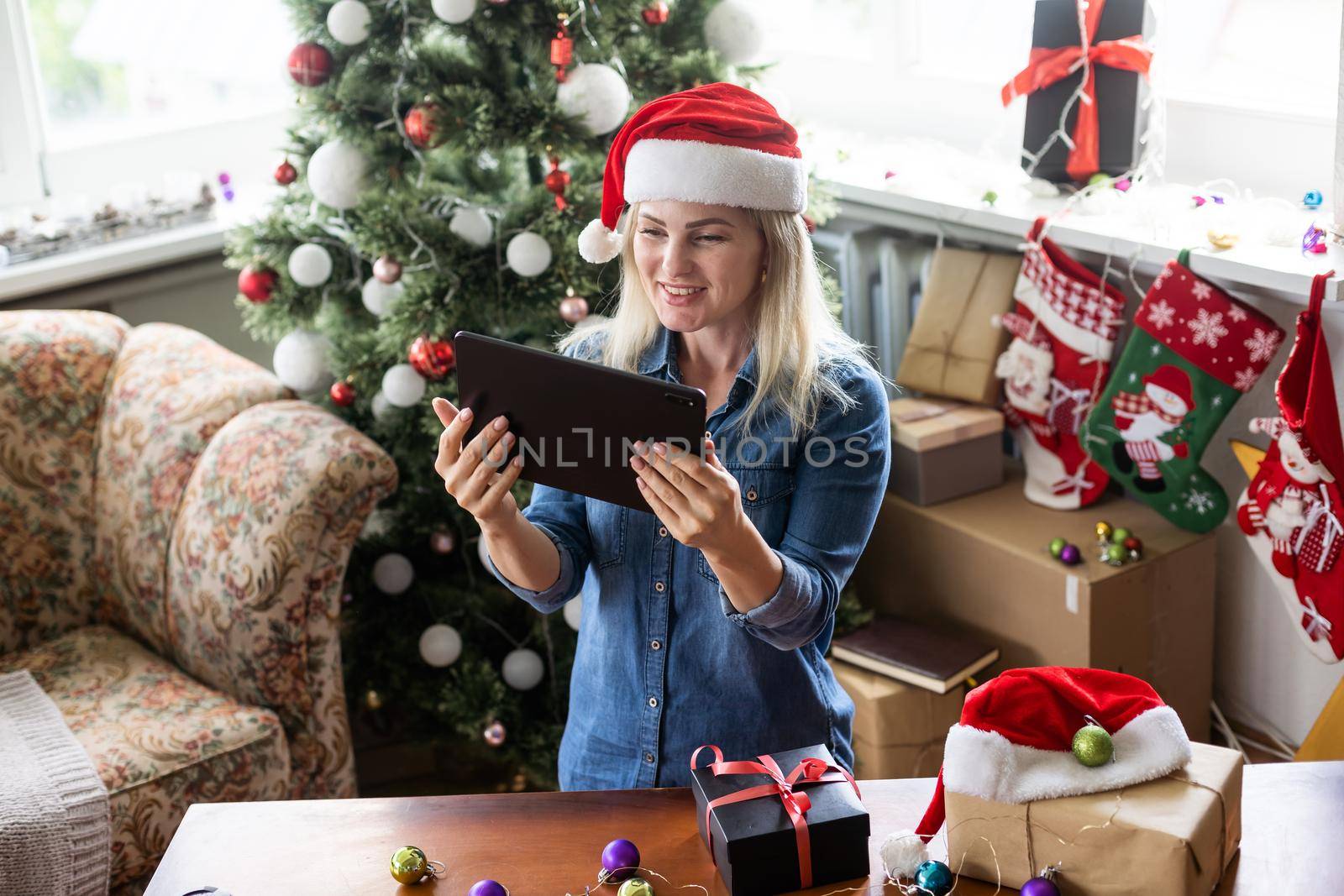 Christmas online holiday. Happy woman having video call with their family or friends. Young woman uses a digital tablet near decorated festive tree at home. Virtual meeting.