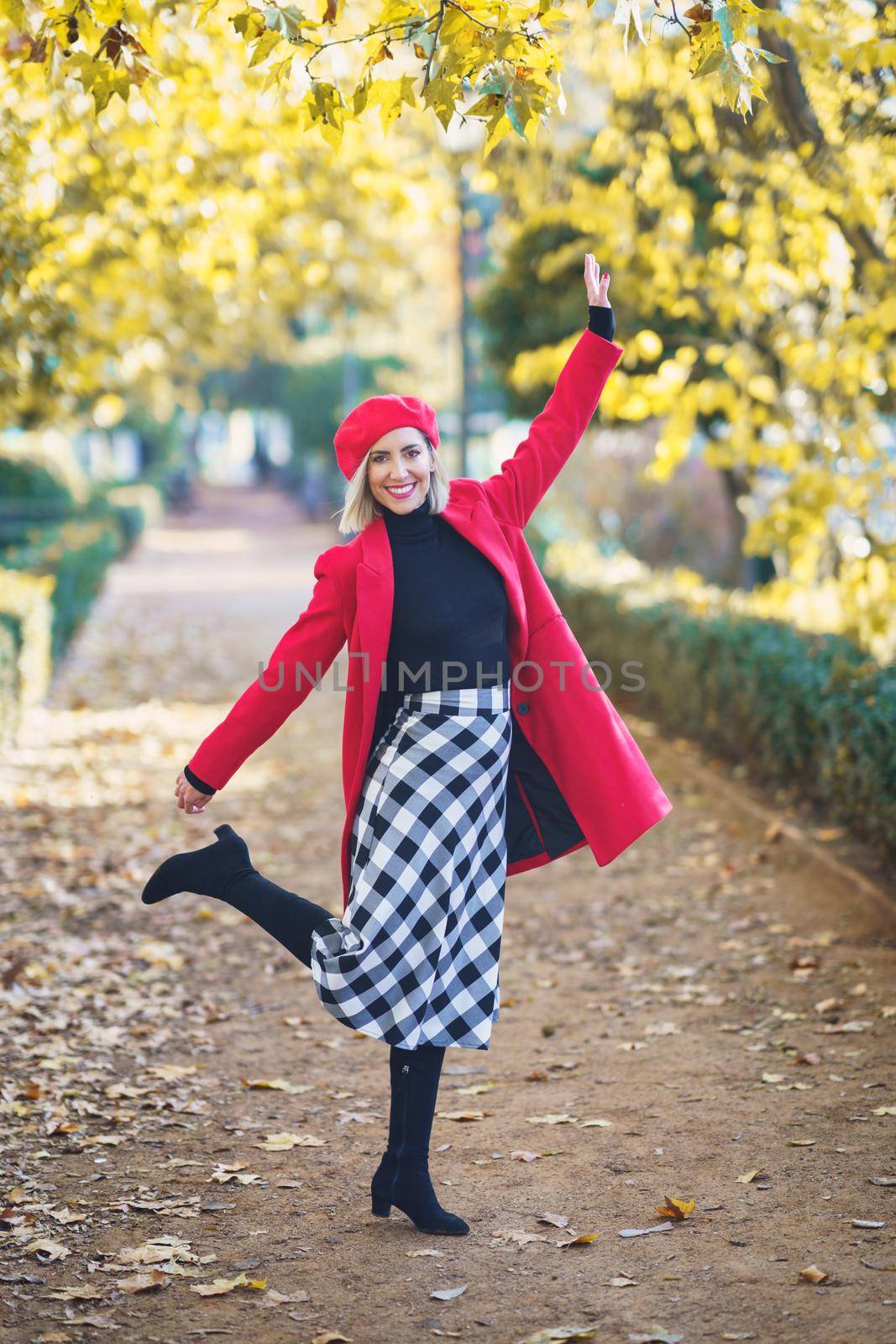 Full body of positive female in red coat and beret, looking at camera with raised arm while standing on path with raised leg in autumn park