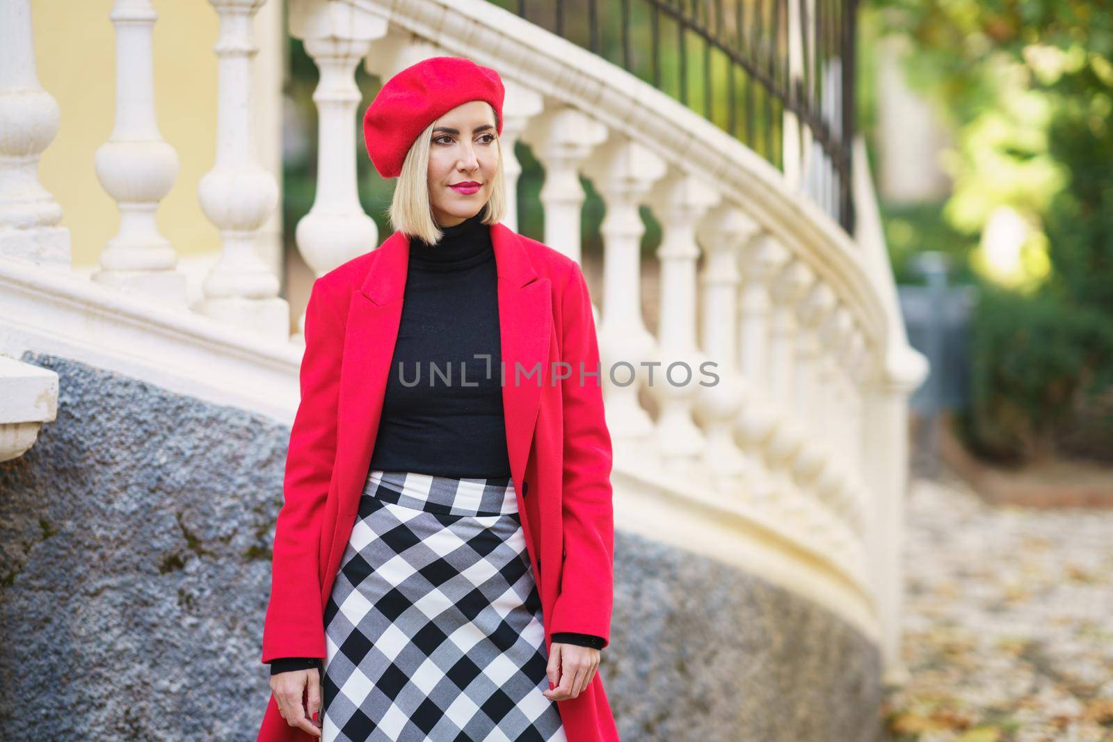 Attractive female in stylish red coat and checkered skirt looking away while standing near white fence on street of city