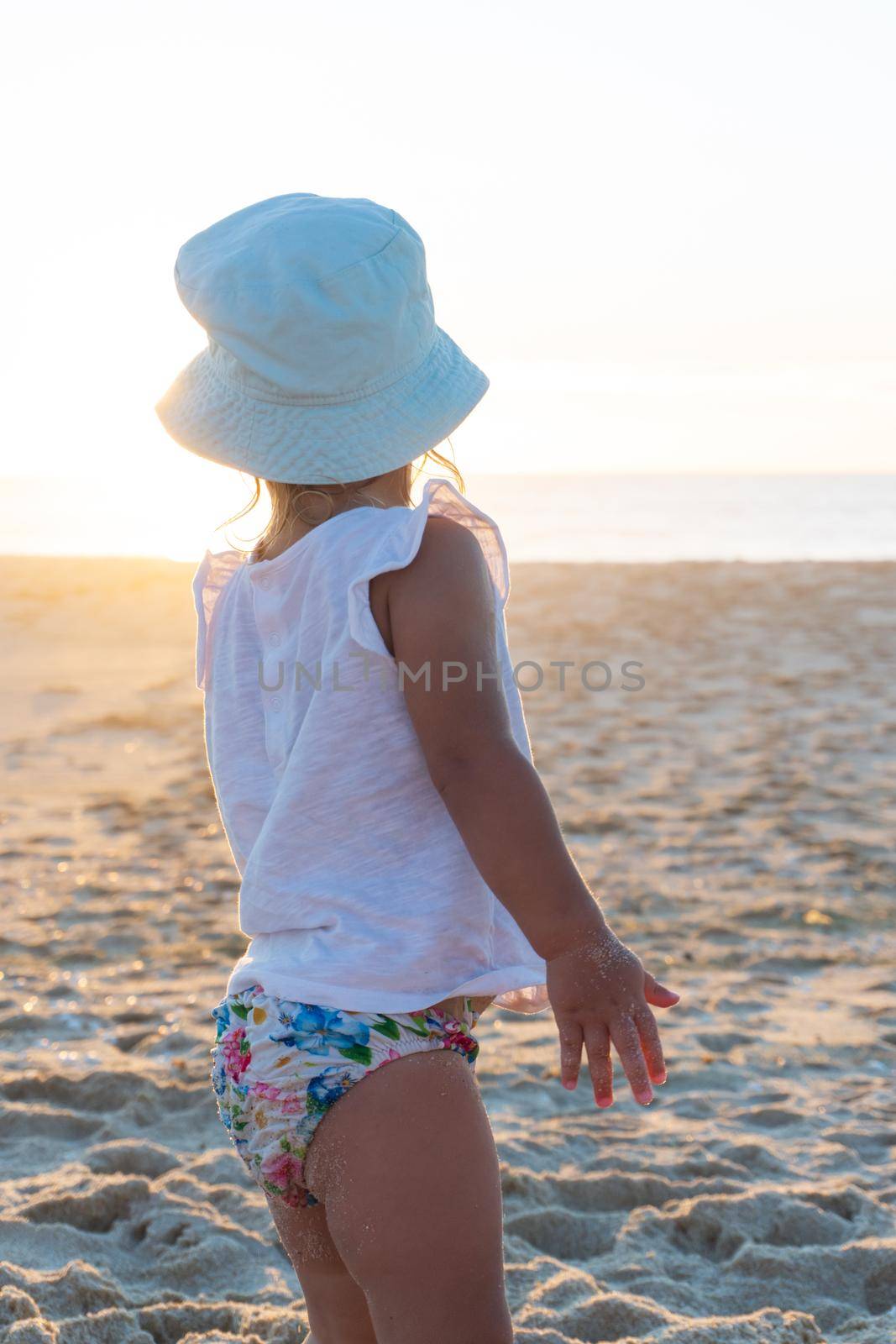 Little girl in a panama hat stands with her back on the beach and looks at the ocean