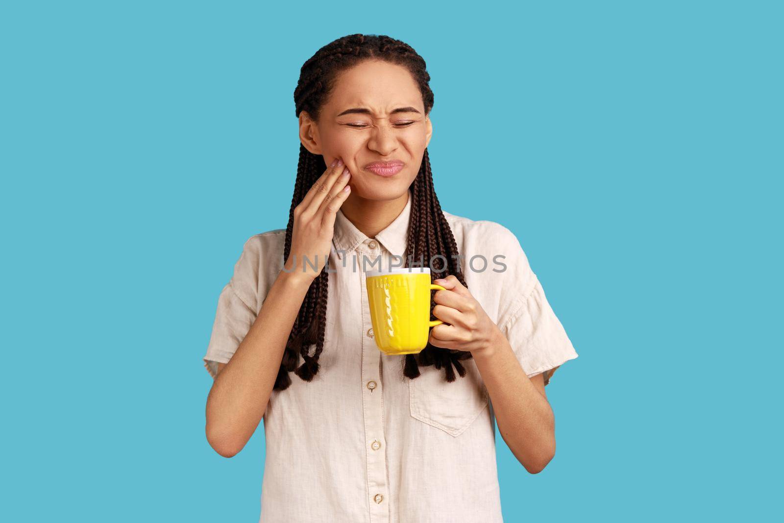 Portrait of unhealthy woman with black dreadlocks wearing white shirt. holding cup in hands, having tooth pain after drinking cold or hot beverage. Indoor studio shot isolated on blue background.