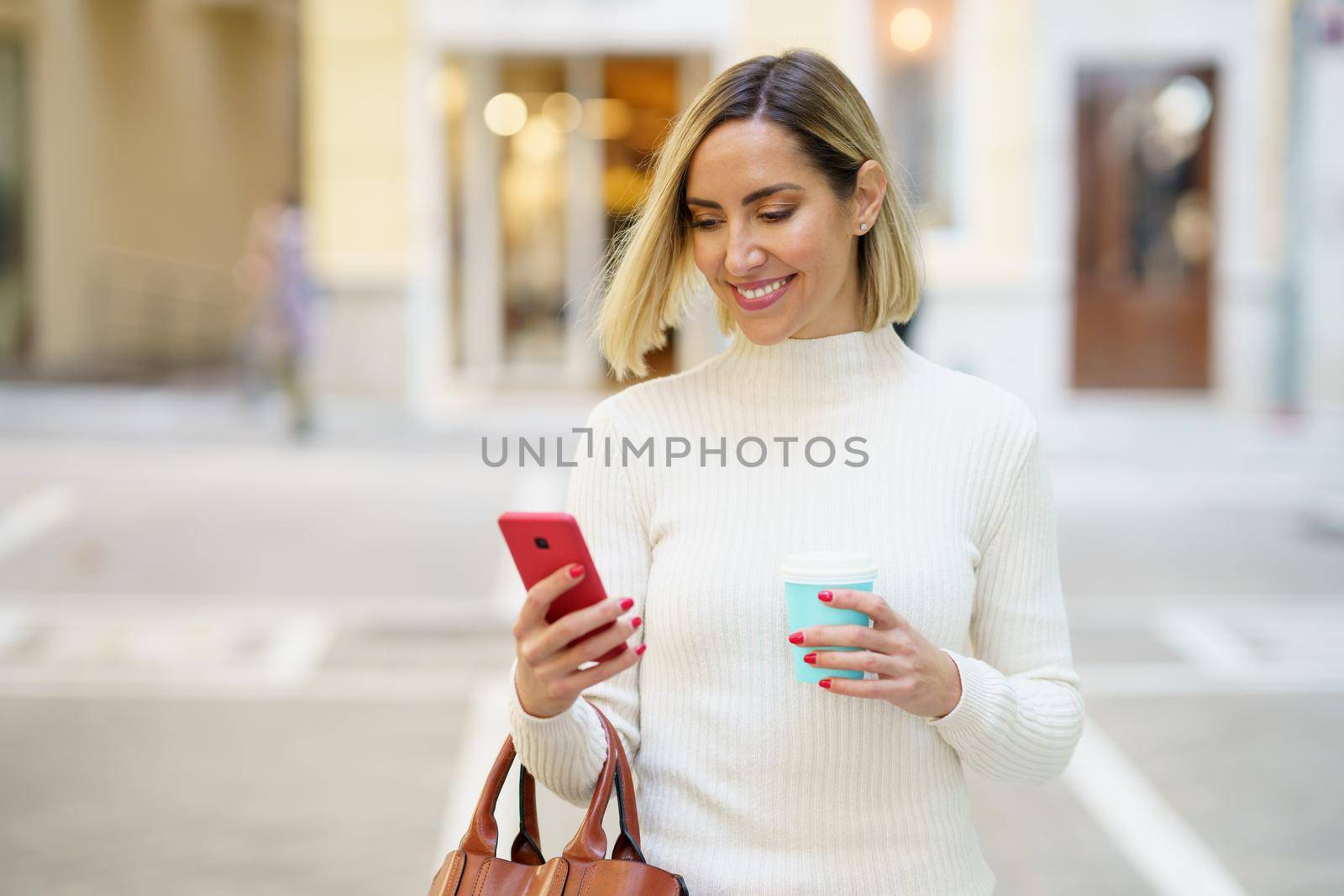 Happy female with blond hair wearing white turtleneck browsing cellphone and holding cup of takeaway beverage