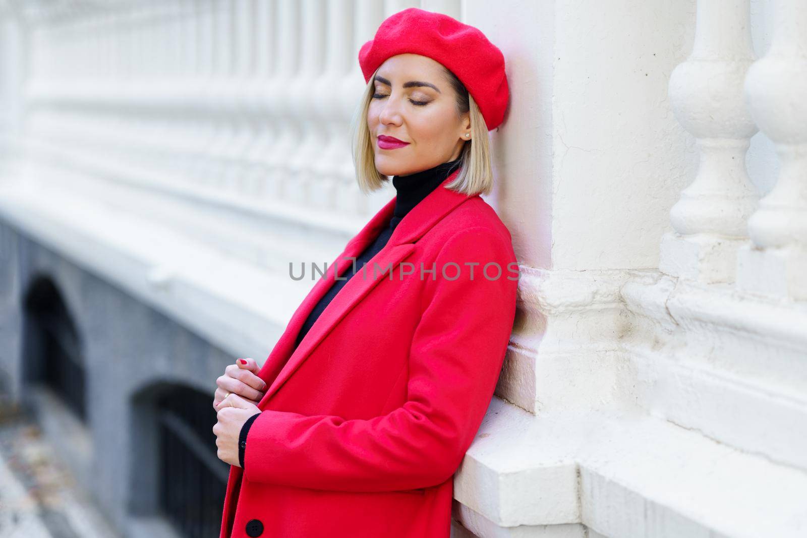 Dreamy female with blond hair in red clothes with beret standing with closed eyes near white fence on street of city