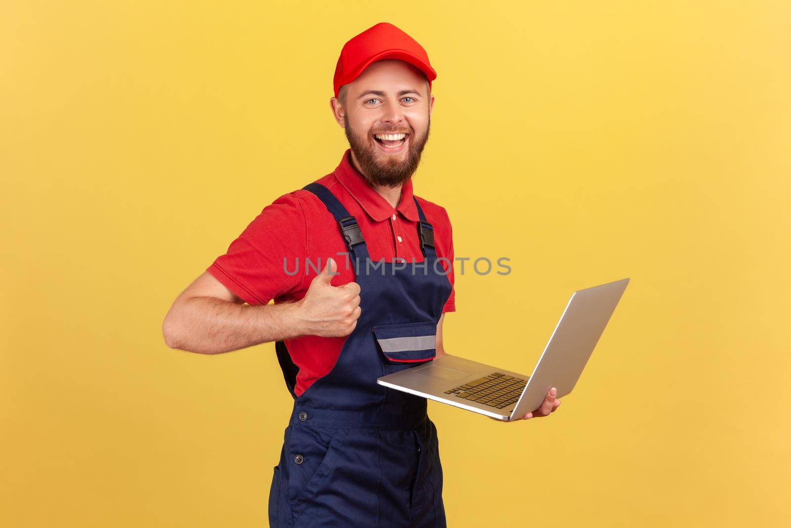 Satisfied worker man wearing blue overalls, red T-shirt and cap working on laptop, showing thumb up, likes a new online service, looking at camera. Indoor studio shot isolated on yellow background.