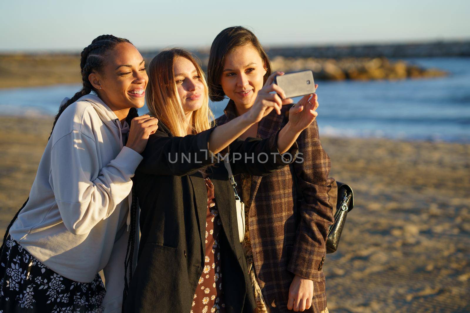 Positive multiethnic female friends taking self portrait on modern smartphone while standing on sandy shore near sea on sunny day